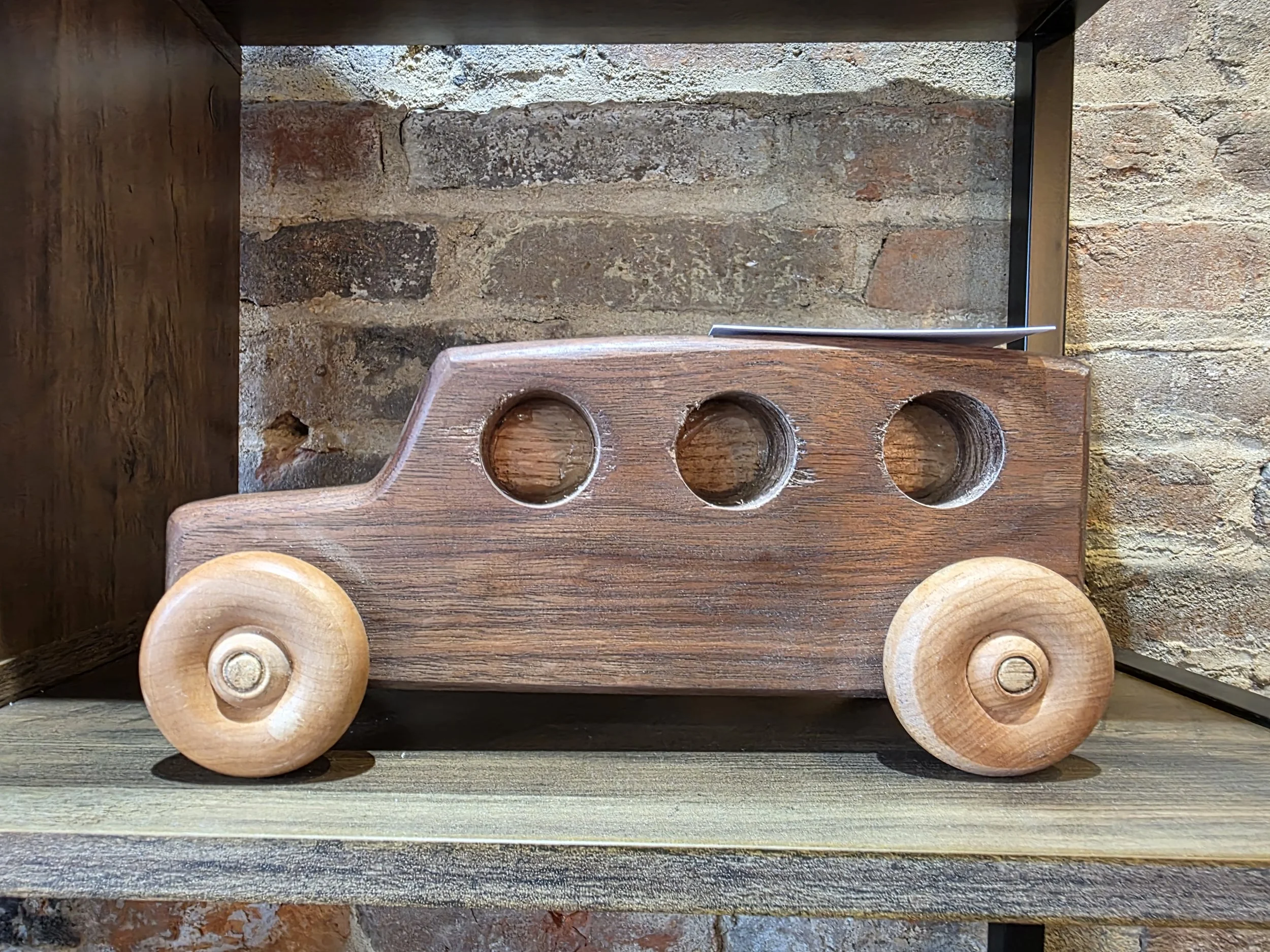 Wooden toy truck with three circular cutouts in the side, light-colored wooden wheels, set against a brick wall background.