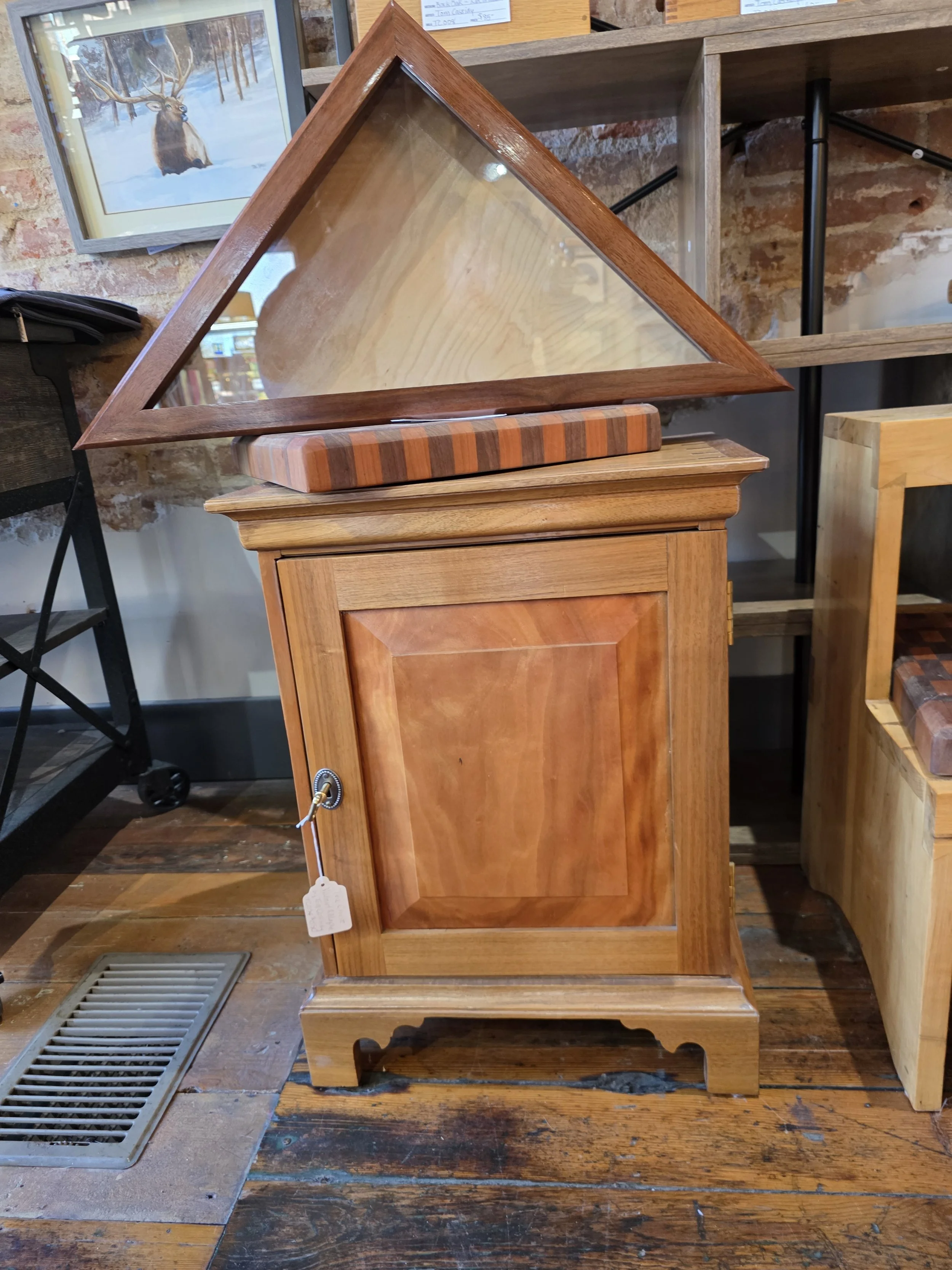 A vintage wooden cabinet with a glass display case on top, featuring a striped cushion. The cabinet is placed on a wooden floor with a brick wall background.