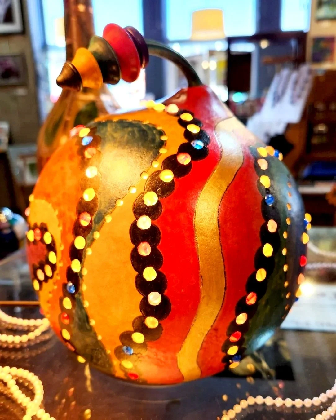 Decorative pumpkin lantern with black, gold, red, and green painted patterns and illuminated dots, surrounded by pearl necklaces, in a cozy indoor setting.