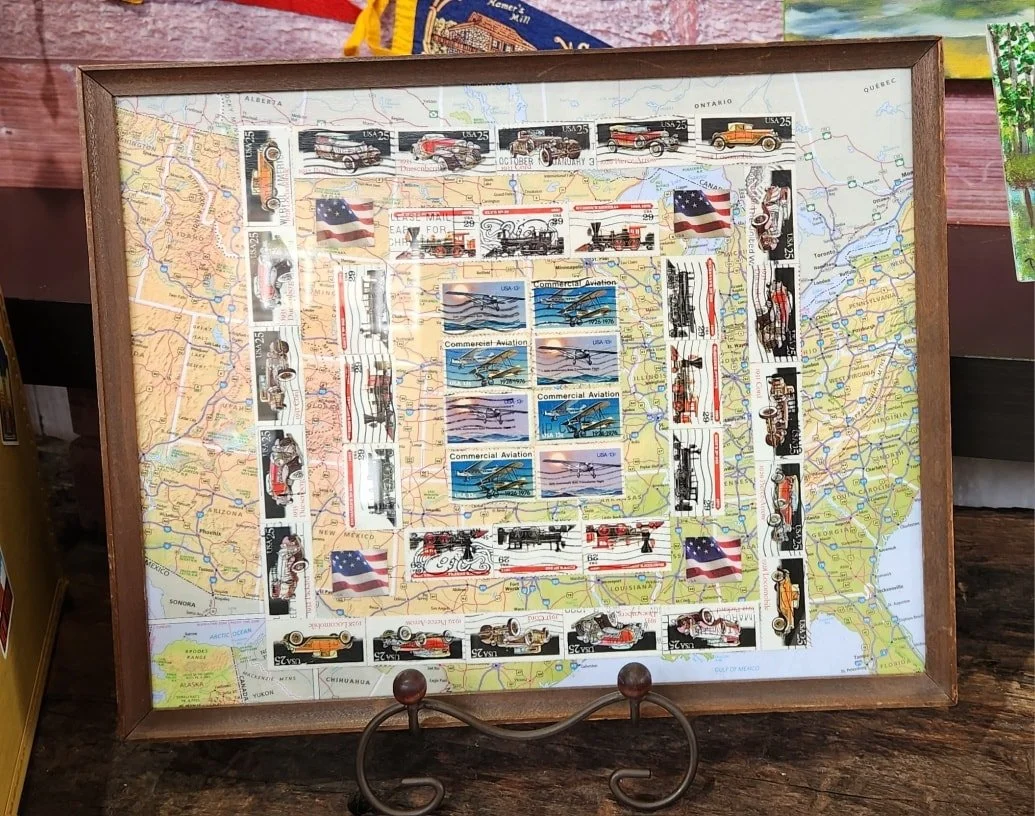 A framed collage of United States stamps arranged on a map of the USA. The stamps feature vintage cars, aircraft, motorcycles, and American flags.