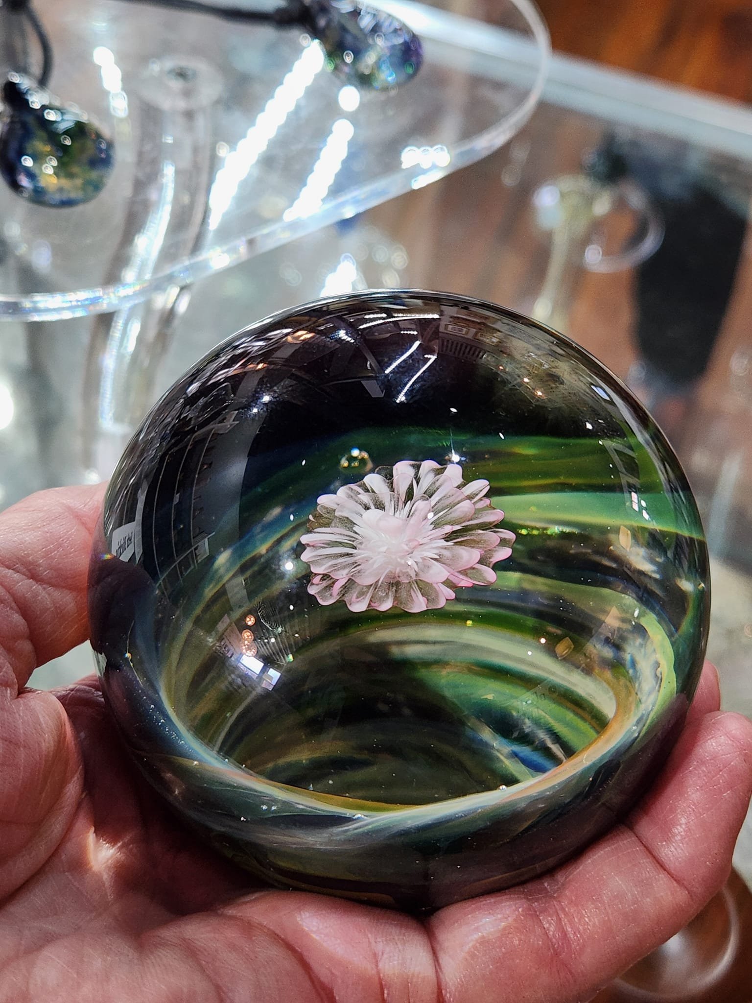 A hand holding a glass paperweight with a pink flower inside, surrounded by swirling green and black patterns.