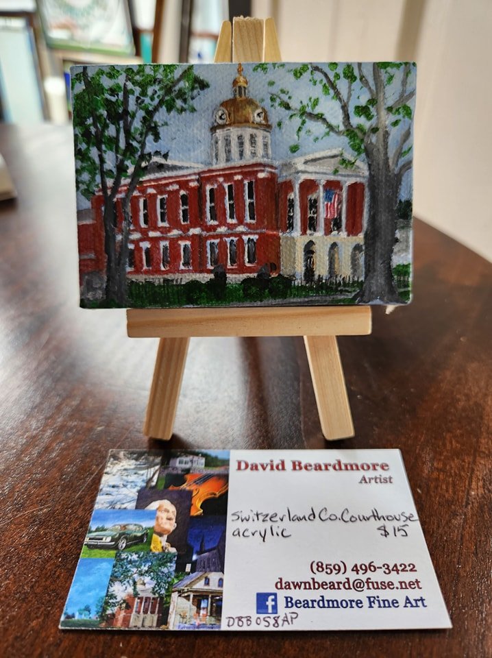 Miniature painting of the Switzerland County Courthouse, an historic red brick building with a clock tower, set on a small easel with a business card below showing artist information.