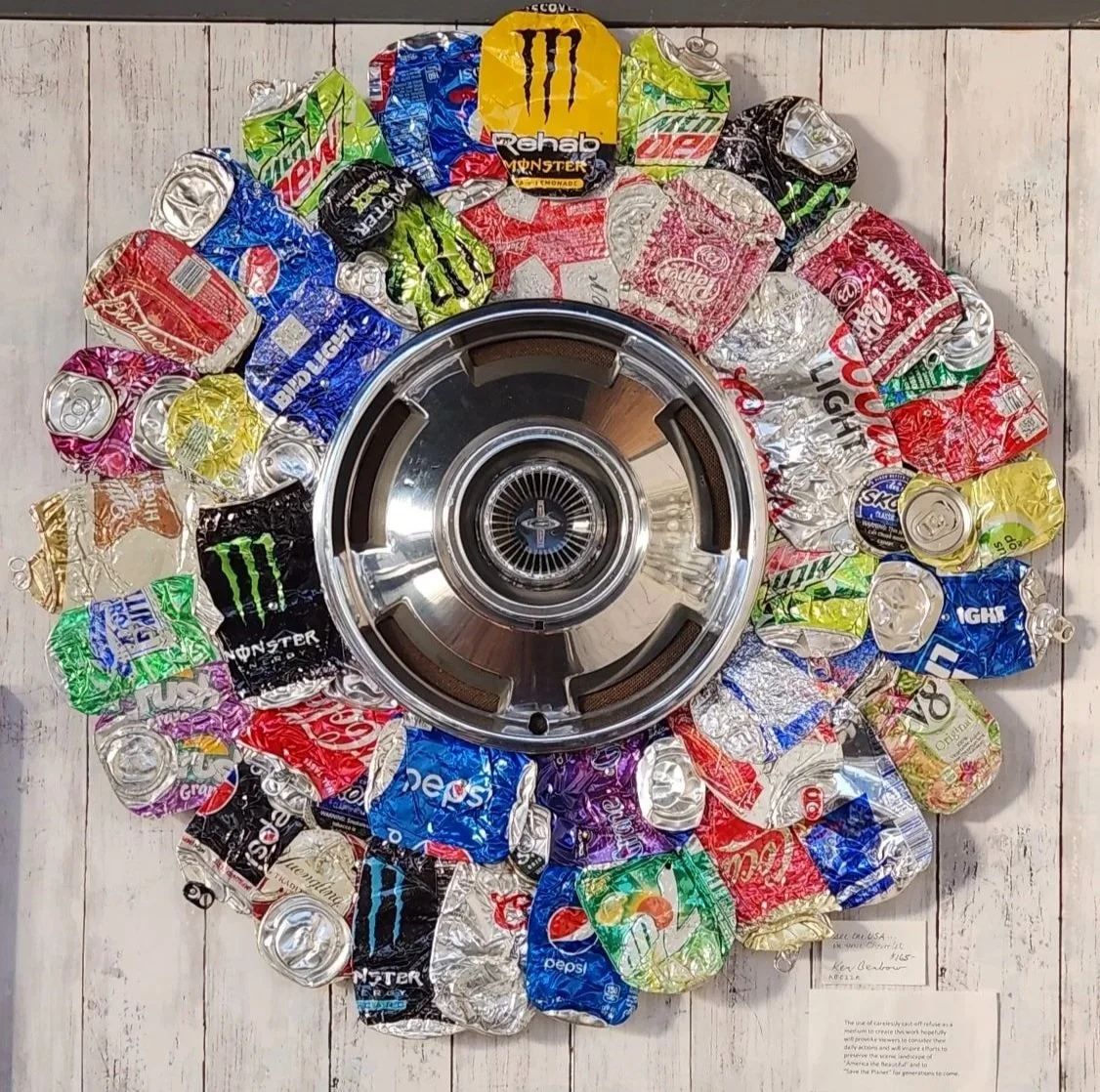 A circular arrangement of crushed soda and energy drink cans around a chrome car wheel on a white wooden background.