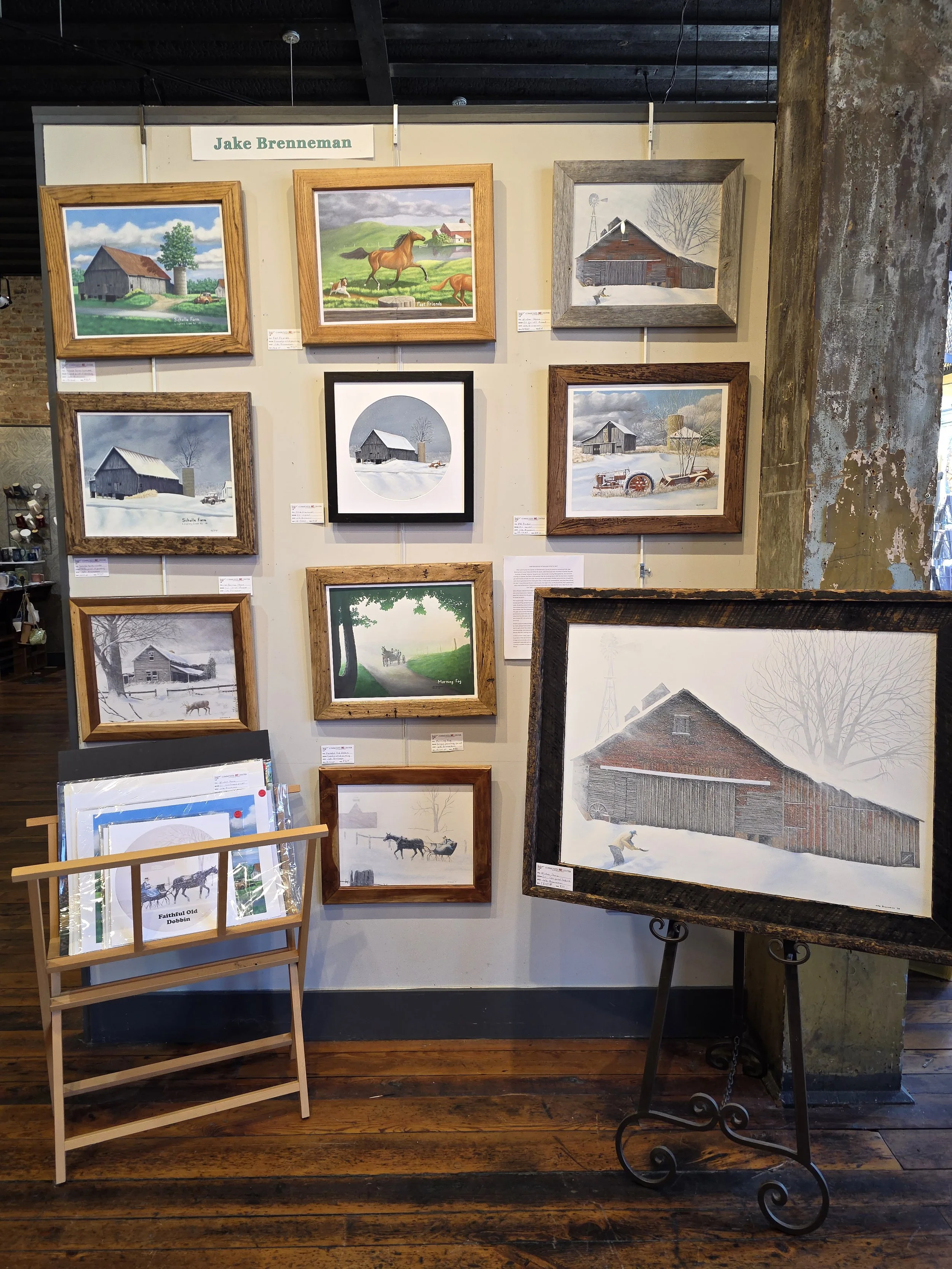 Display of various framed watercolor and oil paintings of rural farm scenes, including barns, horses, and snow-covered landscapes, with a sign labeled 'Jake Brenneman' at the top.