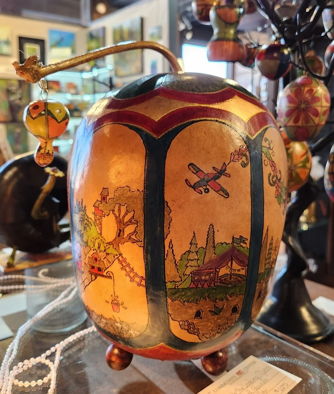 Decorative ceramic vase with painted countryside scene, including a treehouse, an airplane, and a tent, surrounded by hanging painted ornaments in an art store.