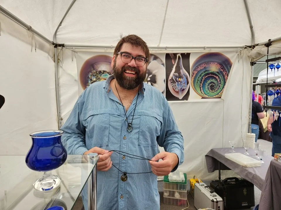 A man with glasses, a beard, and a blue shirt smiling at a booth with art glass pieces, including a blue vase, at an art fair or craft show.