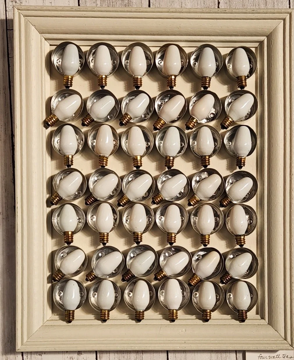 A framed display of multiple vintage style small lightbulbs with white glass and brass fittings arranged in a grid pattern.