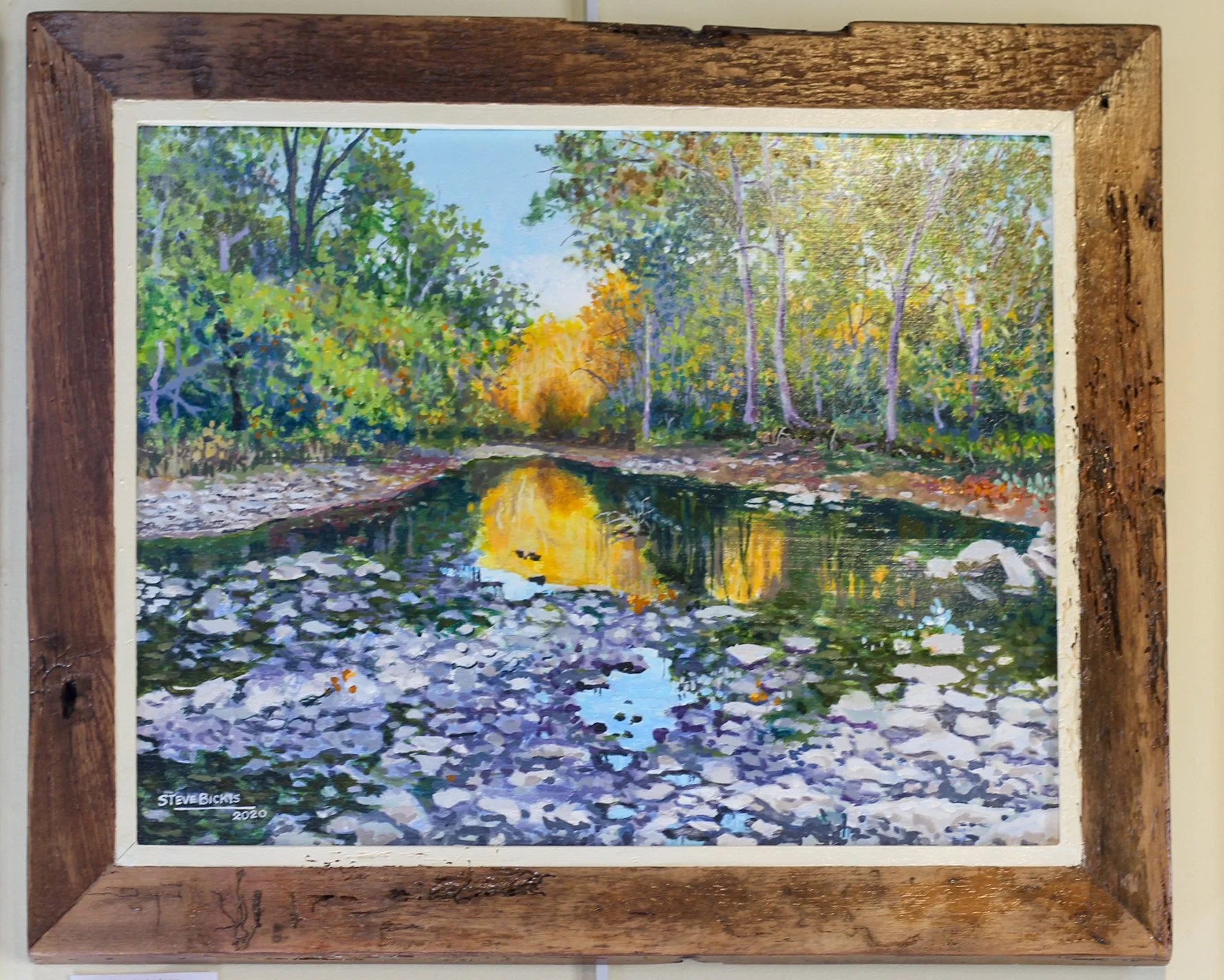 A landscape painting of a river surrounded by trees with autumn foliage, reflecting the sky and trees in the water, framed in rustic wood.