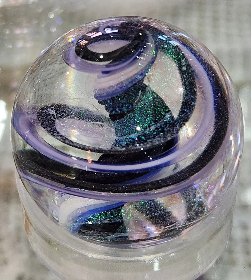 Close-up of a glass paperweight with swirling patterns of purple and black, containing glitter and iridescent elements.