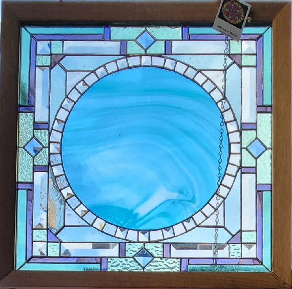A stained glass window with a circular blue design at the center, surrounded by geometric patterns in purple, green, and clear glass, framed in wood.