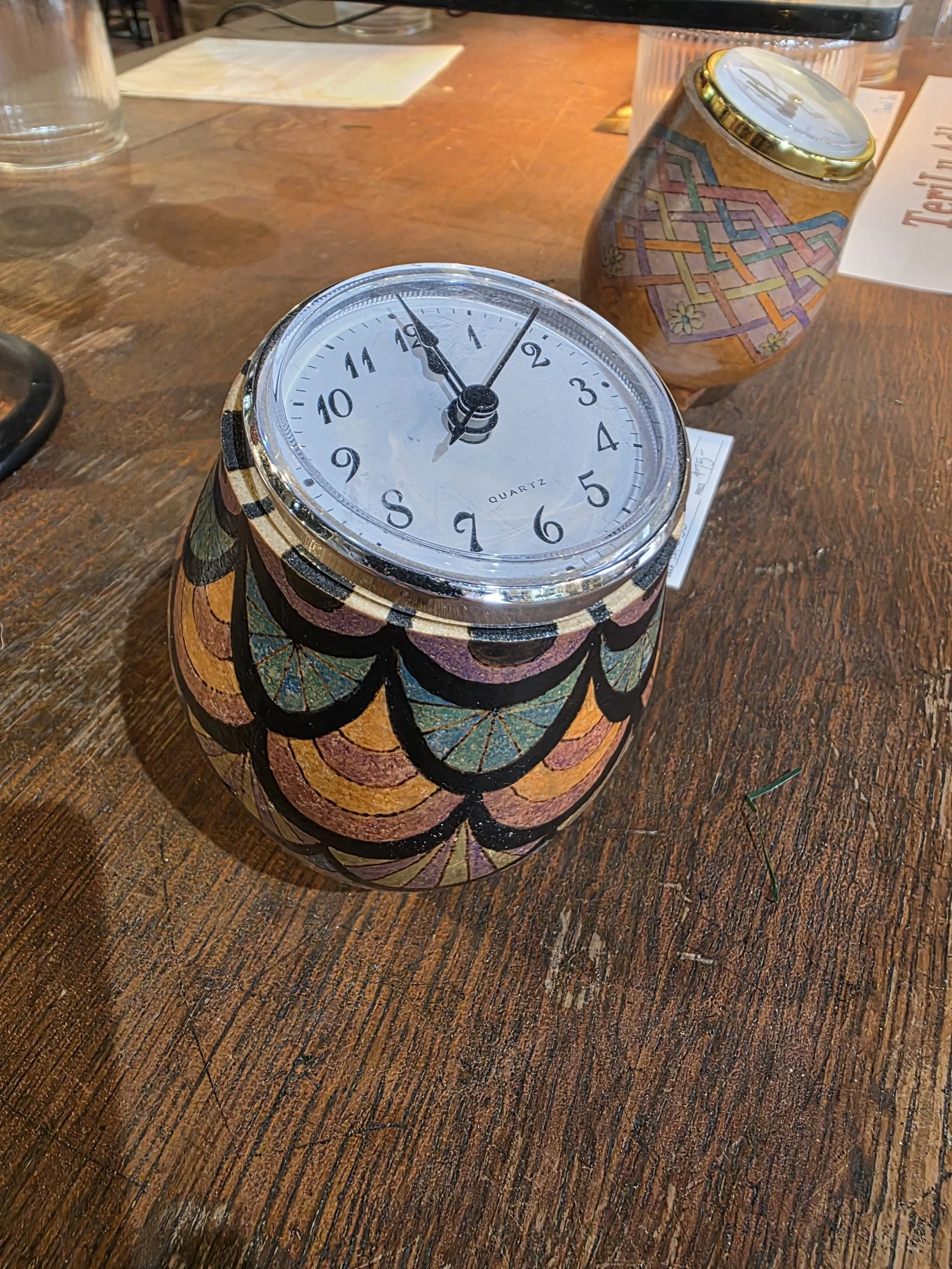 Decorative clock with a colorful, patterned base on a wooden table, showing the time as approximately 11:56.