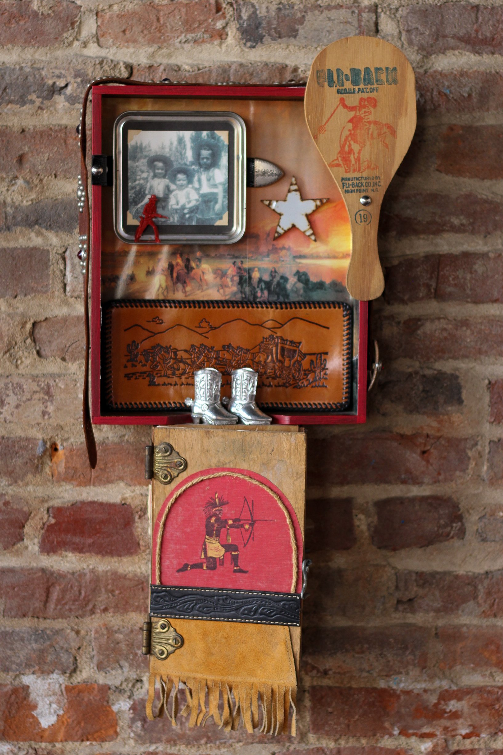 Decorative display on a brick wall including a small photo of three children, a wooden paddle with a cow and rider, a vintage cowgirl badge, miniature silver cowboy boots, a western-themed leather case, and a fringed leather pouch with a painted Native American figure.