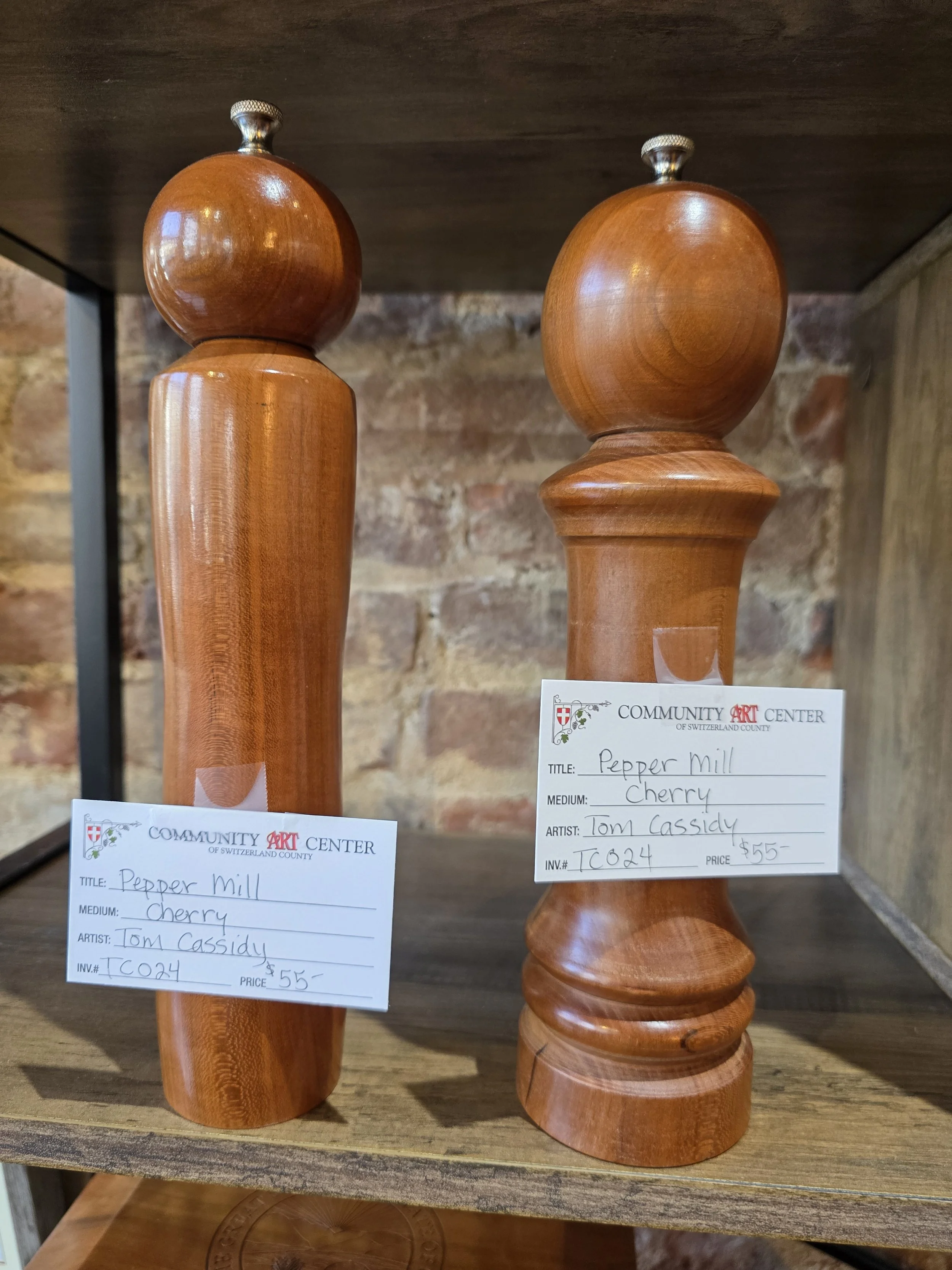 Two wooden pepper mills on a wooden shelf, each with a white label from the Community Art Center. The labels indicate they are cherry wood, crafted by Tom Cassidy, priced at $55.