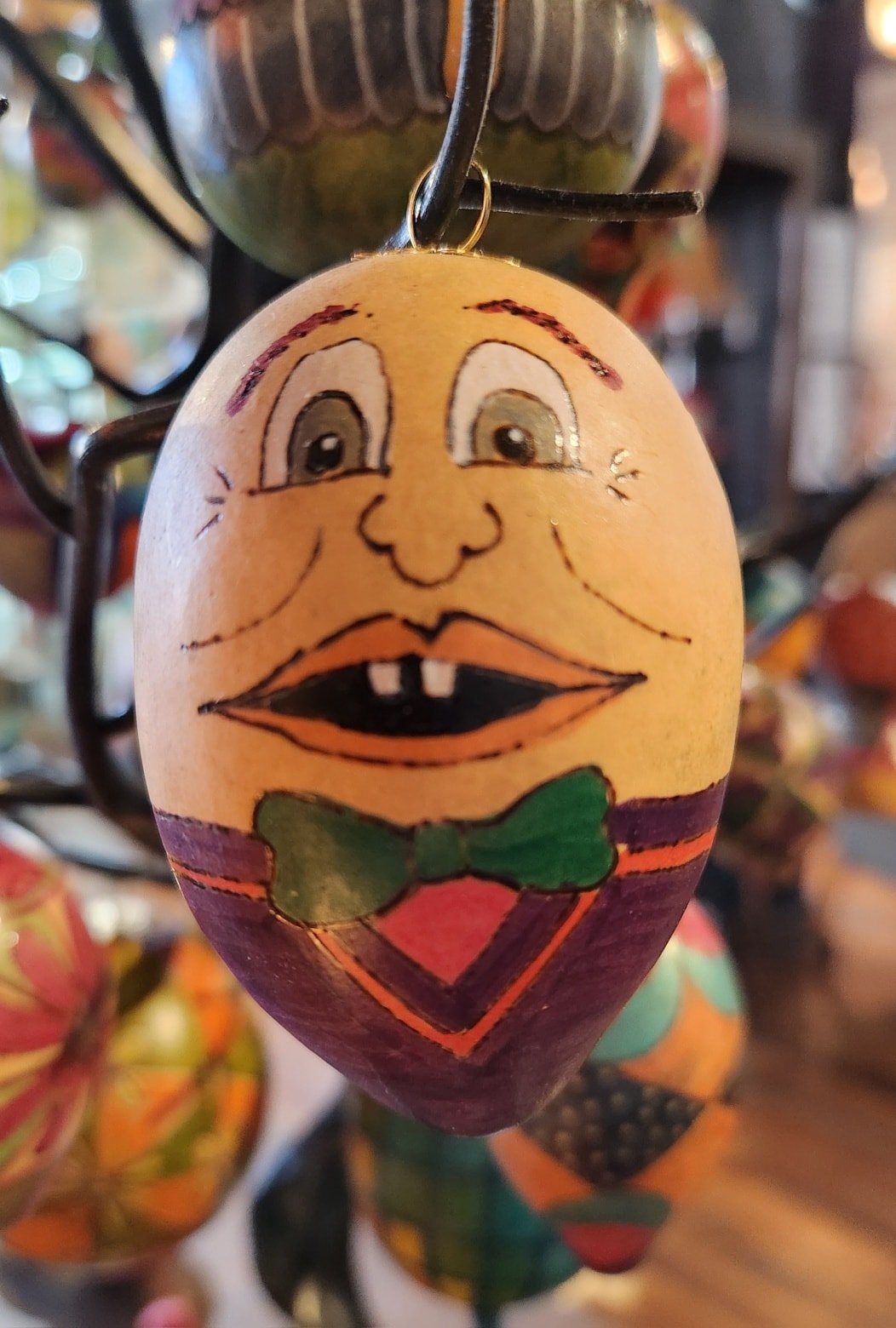A painted decorative egg with a cartoon face, showing big eyes, a wide smiling mouth with two front teeth, and a green bow tie, hanging among other colorful painted eggs.