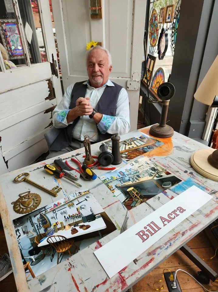 An elderly man with gray hair and beard, dressed in a checkered shirt and gray vest, sitting at a cluttered table inside an art studio. The table has various tools, artifacts, and photos, with a sign reading 'Bill Acra' in front of him. Bright, colorful artwork and crafts are visible in the background.