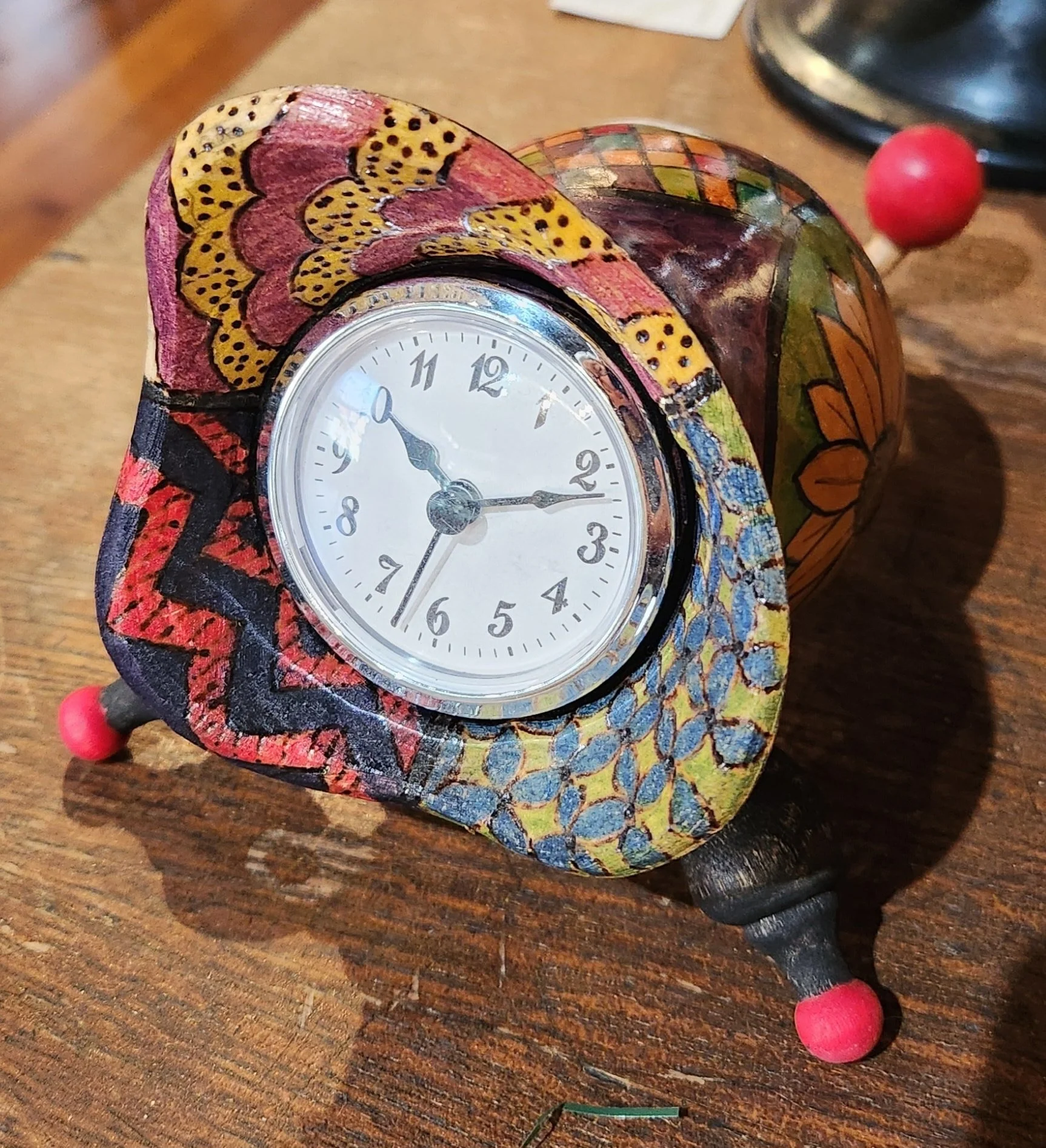 A small, colorful, hand-painted clock with a round face, set on a wooden surface. The clock has a floral and geometric design with pink, yellow, blue, and black colors, and red feet.
