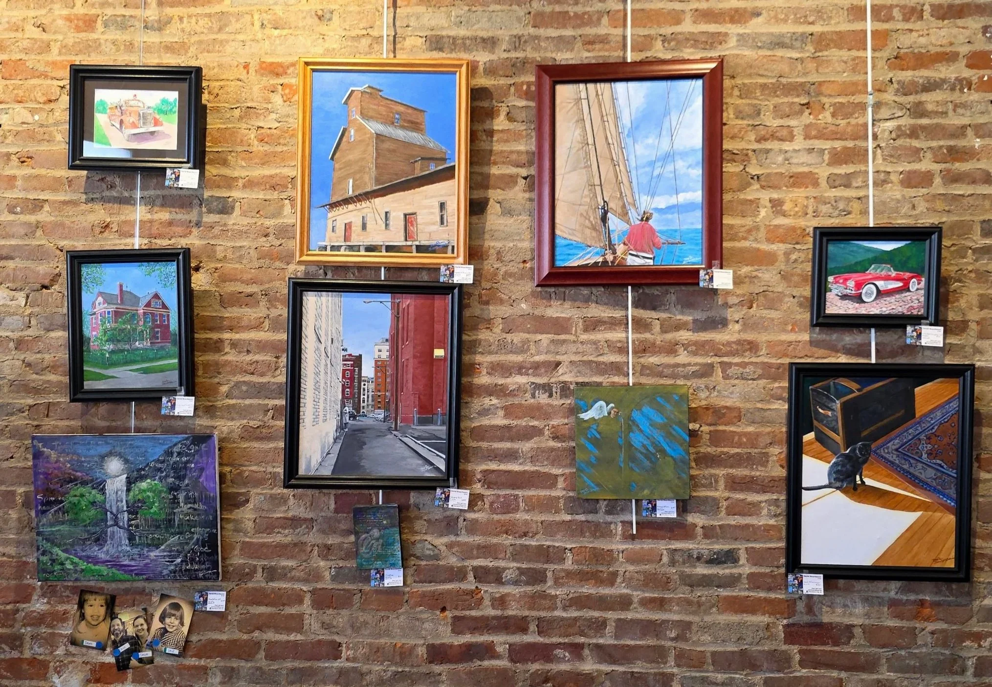 An art gallery wall decorated with various paintings and photographs, including a house, a building, a sailboat, a car, a street scene, a forest scene, a bird, and children’s photos, all mounted on a brick wall.