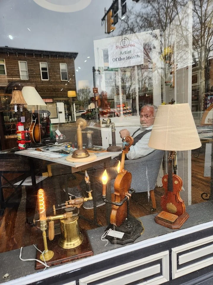 View through the shop window showing a man sitting at a desk surrounded by antique items, including lamps, a guitar, a brass lamp with multiple bulbs, and a small ceramic vase. A sign inside highlights 'Artist of the Month.' The street outside has buildings and trees, and the scene reflects in the glass window.