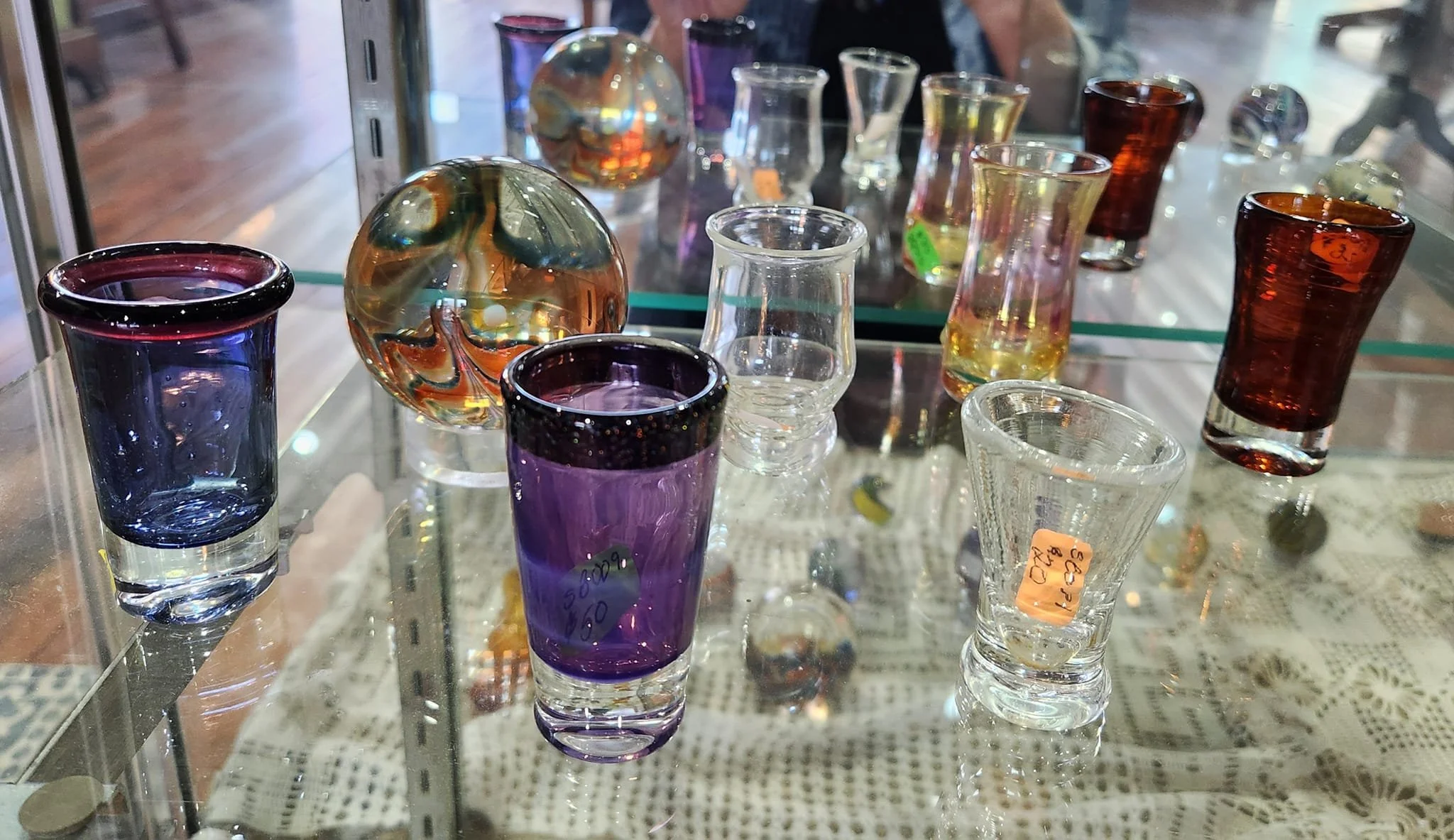 Various colorful glass vases and paperweights displayed on a glass shelf in a store, reflected in the mirror behind.