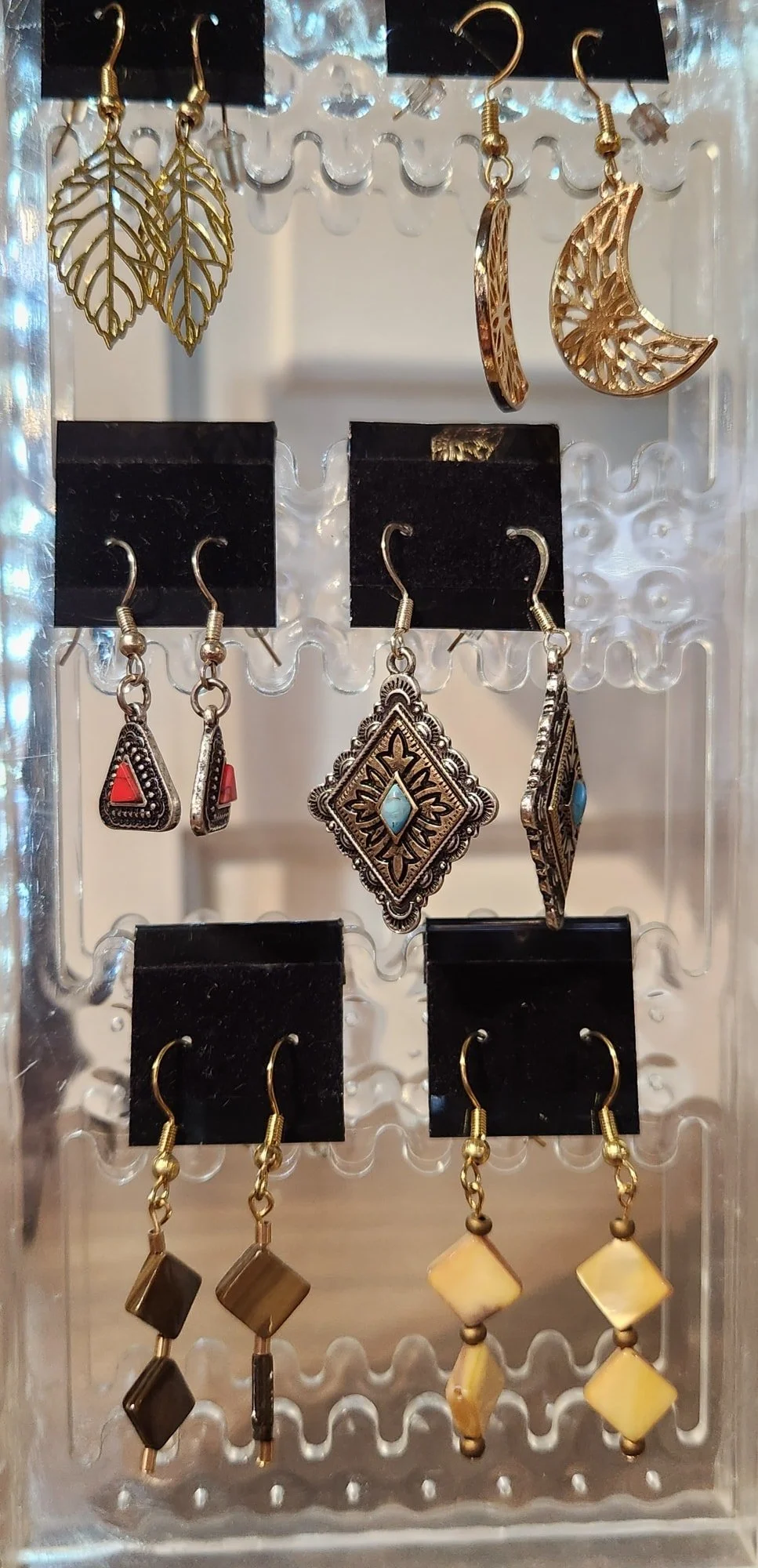 A display case with six pairs of earrings, including gold leaf-shaped earrings, crescent moon earrings, red and turquoise stone earrings, and geometric stone earrings.