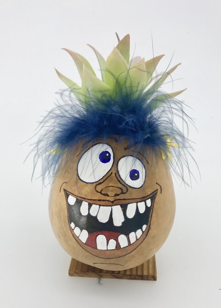 Decorative gourd painted with a cartoon face, featuring large eyes, a wide open mouth with big teeth, blue wig-like hair, and pineapple leaves on top, mounted on a wooden base.