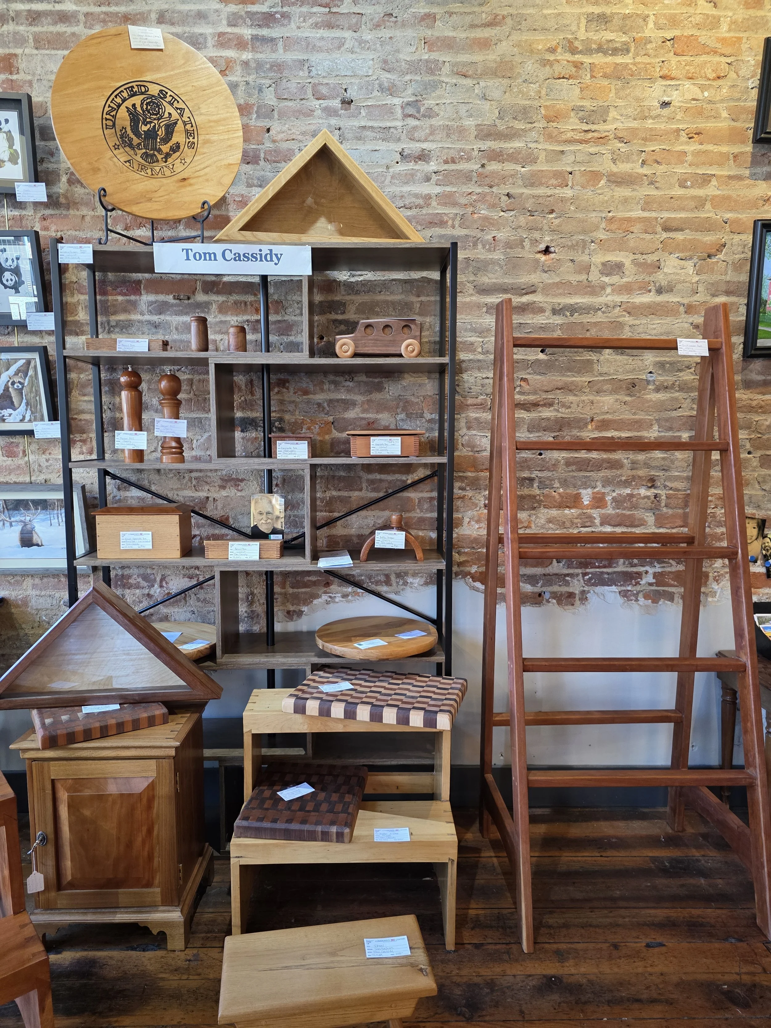 Shelf with wooden and fabric furniture and objects, including a ladder, display cases, and framed pictures, against a brick wall with a sign reading 'Tom Cassidy'.