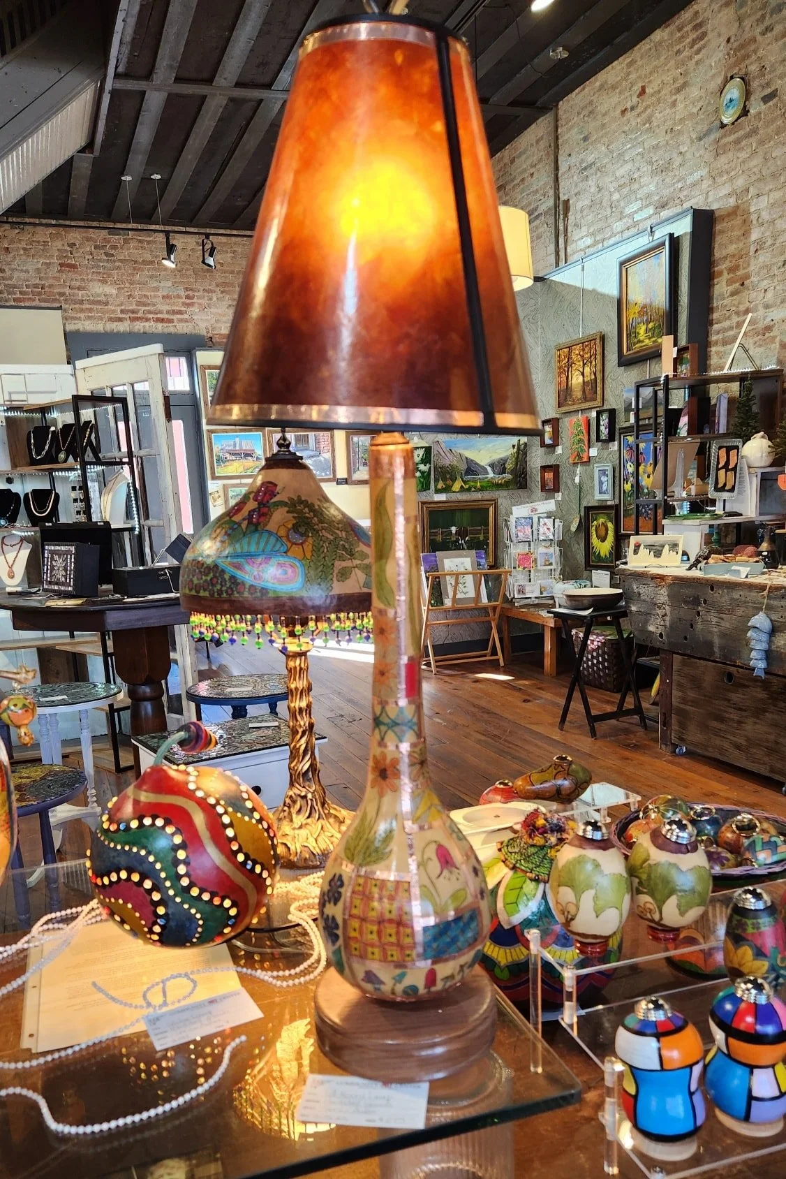Decorative lamps and colorful pottery displayed on glass shelves inside an art gallery or boutique shop with brick walls and framed paintings.