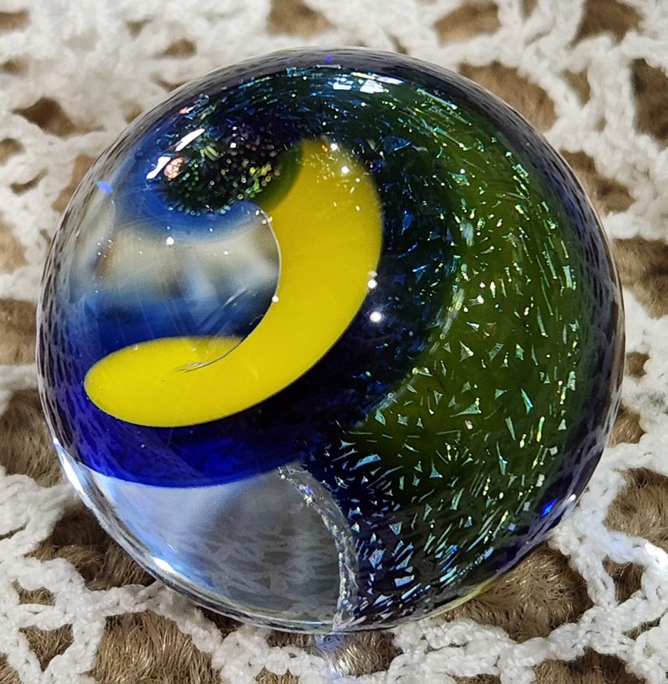Colorful glass paperweight with a yellow, blue, and green swirl design, resting on a woven surface.