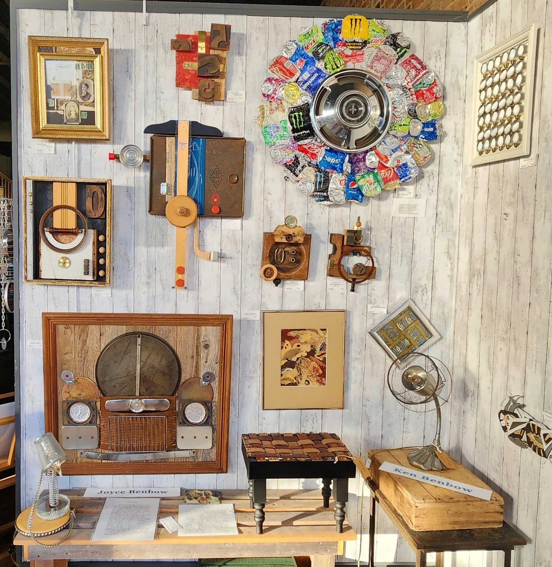 A wall decorated with various vintage and modern art pieces, including framed pictures, sculptures, and a large circular arrangement of crushed aluminum cans around a metal object, with additional objects like drawers, a fan, and artwork displayed on tables below.