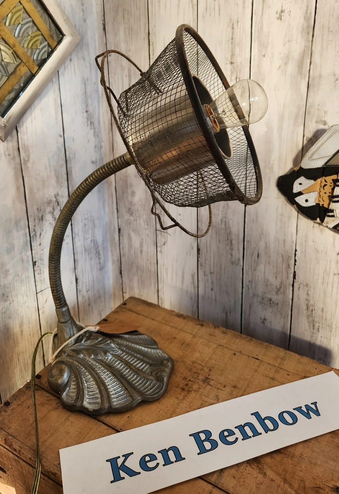 Vintage desk lamp with a wire mesh shade and an Edison-style light bulb on a wooden surface, with a sign reading 'Ken Benbow' in front.