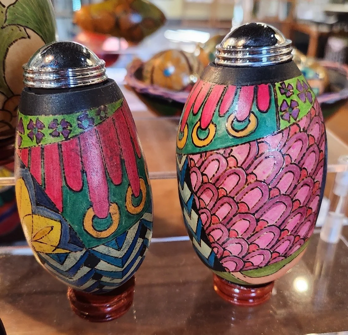 Colorful decorative egg-shaped containers with black openings at the top, painted with pink, green, yellow, blue, and purple patterns, displayed on a wooden surface.