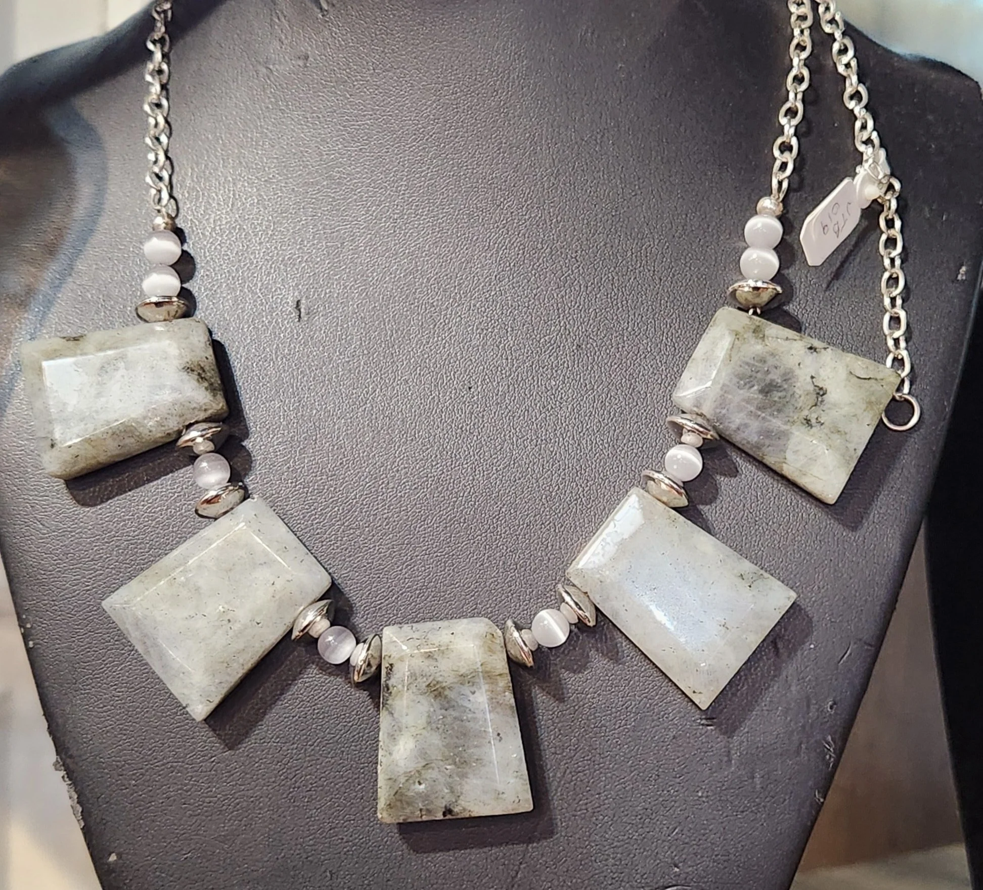 A statement necklace featuring large rectangular white stones, small white beads, and silver beads on a silver chain displayed on a black stand.