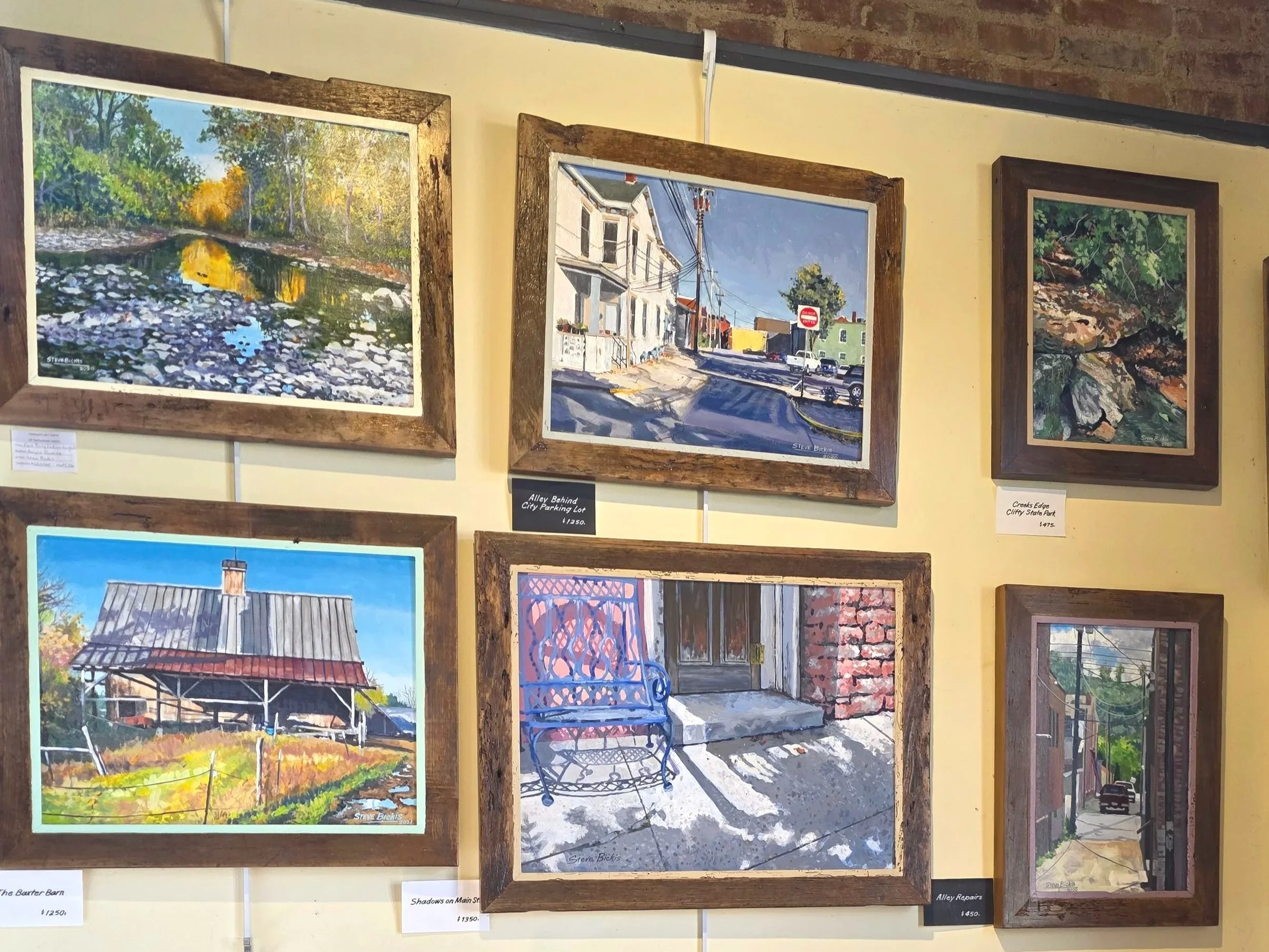 A collection of six landscape paintings displayed on a beige wall in a gallery, framed with wood. The paintings depict outdoors scenes including a river, a street view with utility poles, a creek, a barn, a porch with a bench, and an alley with cars.