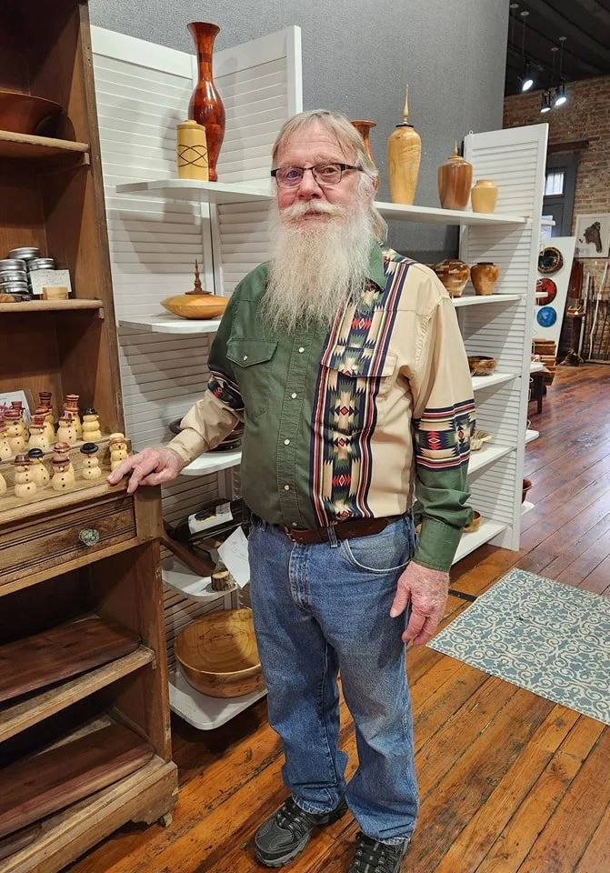 An elderly man with glasses, a long white beard, and long white hair, wearing a patchwork shirt with green and beige colors, blue jeans, and sneakers, standing in a craft store surrounded by wooden pottery and decorative items.