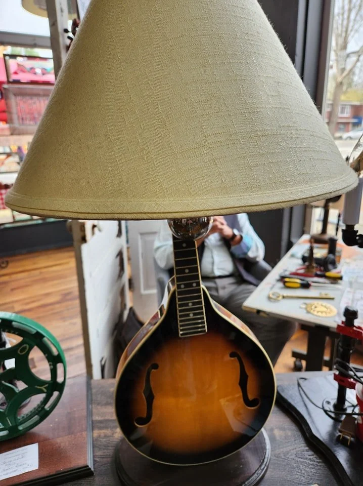 A lamp with a shade on top and a body resembling a guitar, placed on a table in a room with various tools and objects around.