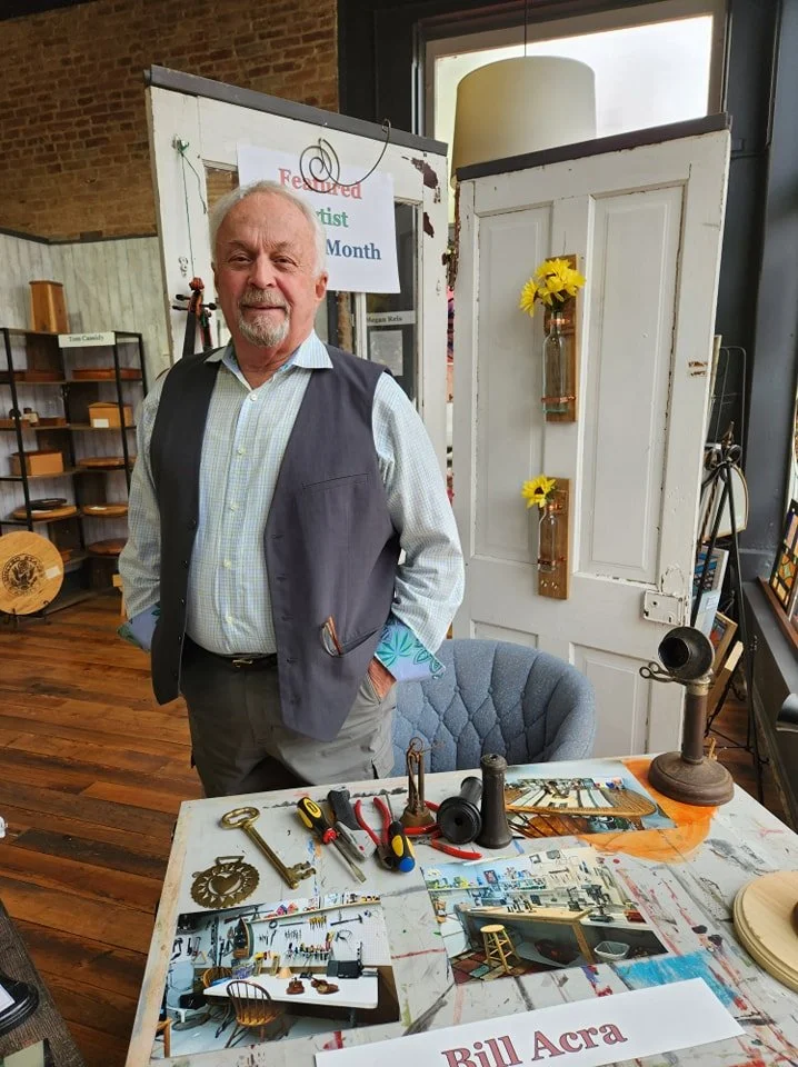 A man standing behind a table with tools and craft supplies in a room with wooden floors, a brick wall, and decorated with yellow flowers in glass bottles. There is a sign behind him that reads 'Featured Artist of the Month'.