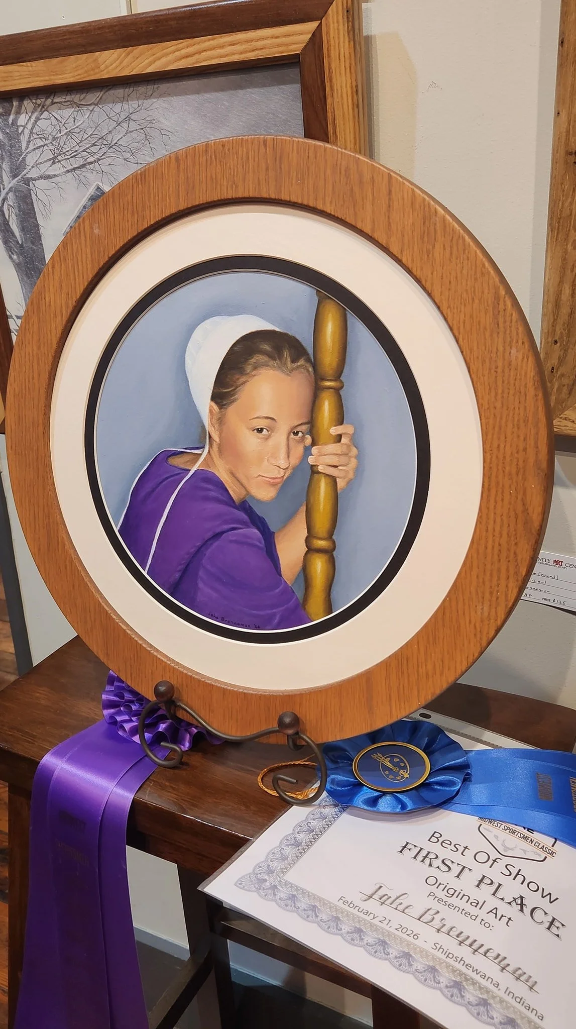 Framed portrait of a woman with light skin and brown hair, wearing a purple shirt and a white hair cap, holding a wooden stick, displayed on a table with purple ribbons and an award certificate for 'Best of Show First Place' presented to Jake Breunig, dated February 21, 2026.