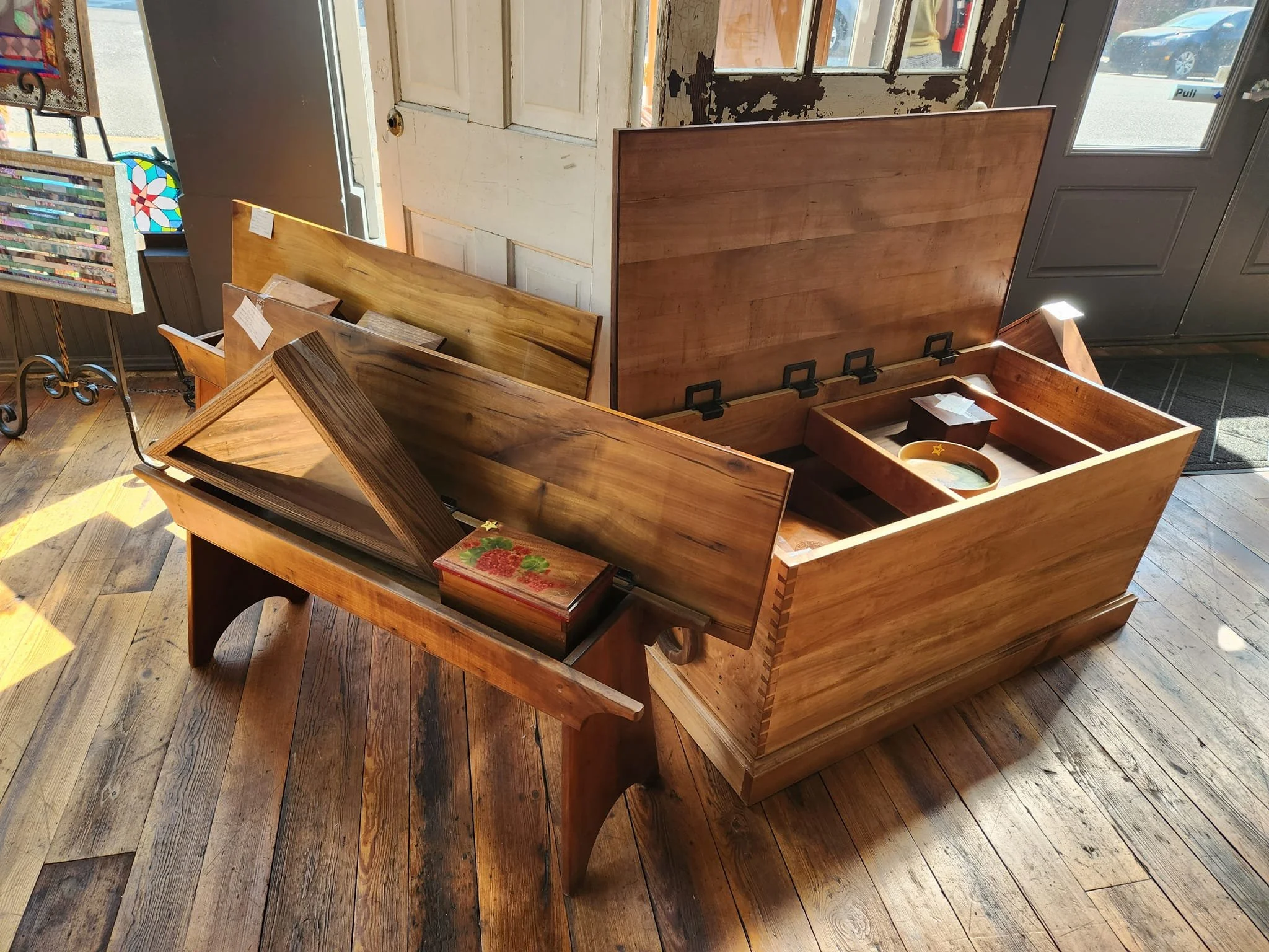 Wooden display case and small wooden bench placed on a wooden floor inside a store with sunlight shining through the windows.