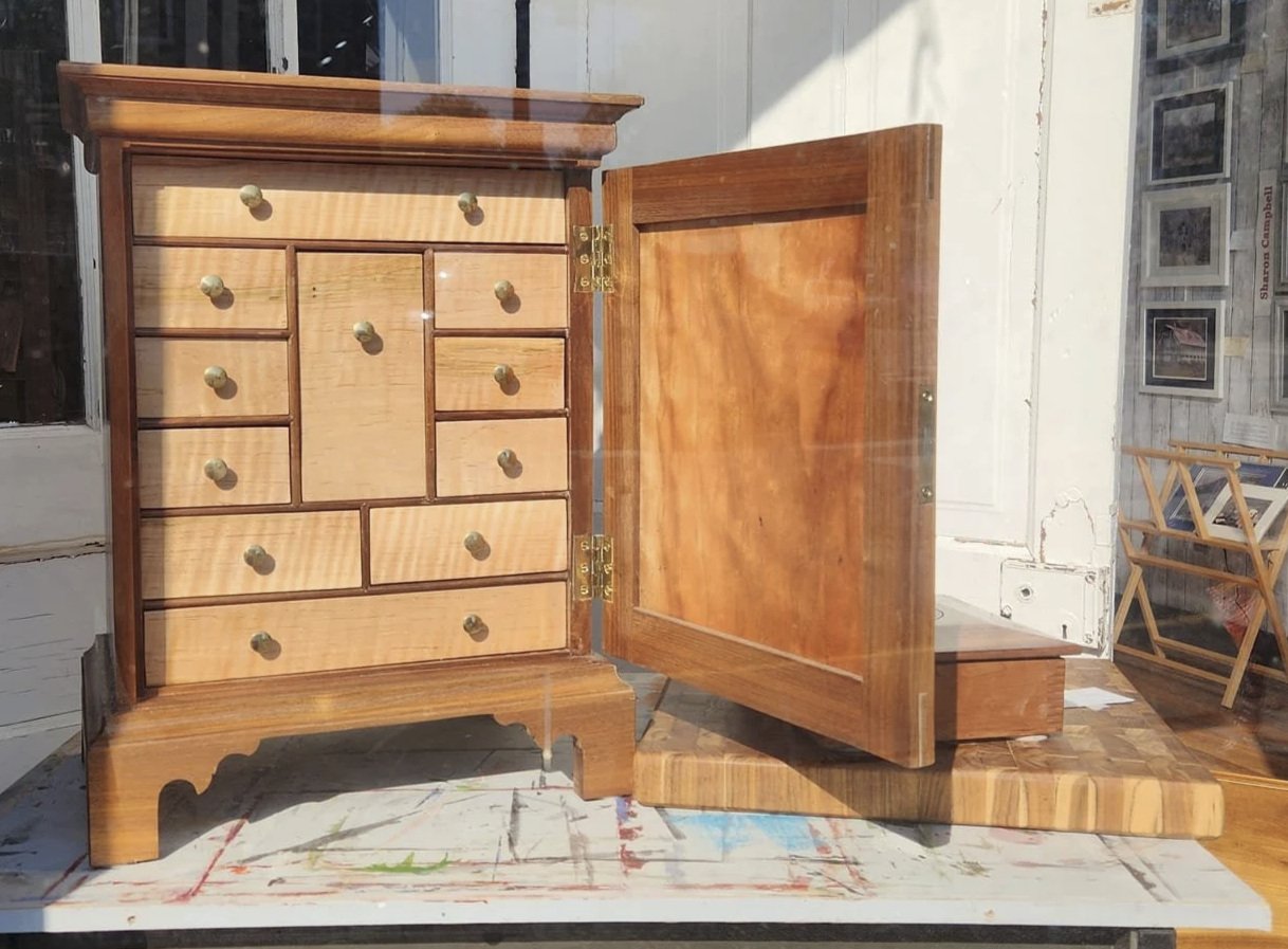 A wooden jewelry box with multiple small drawers and a larger central compartment, placed on a work table in a workshop or studio setting.