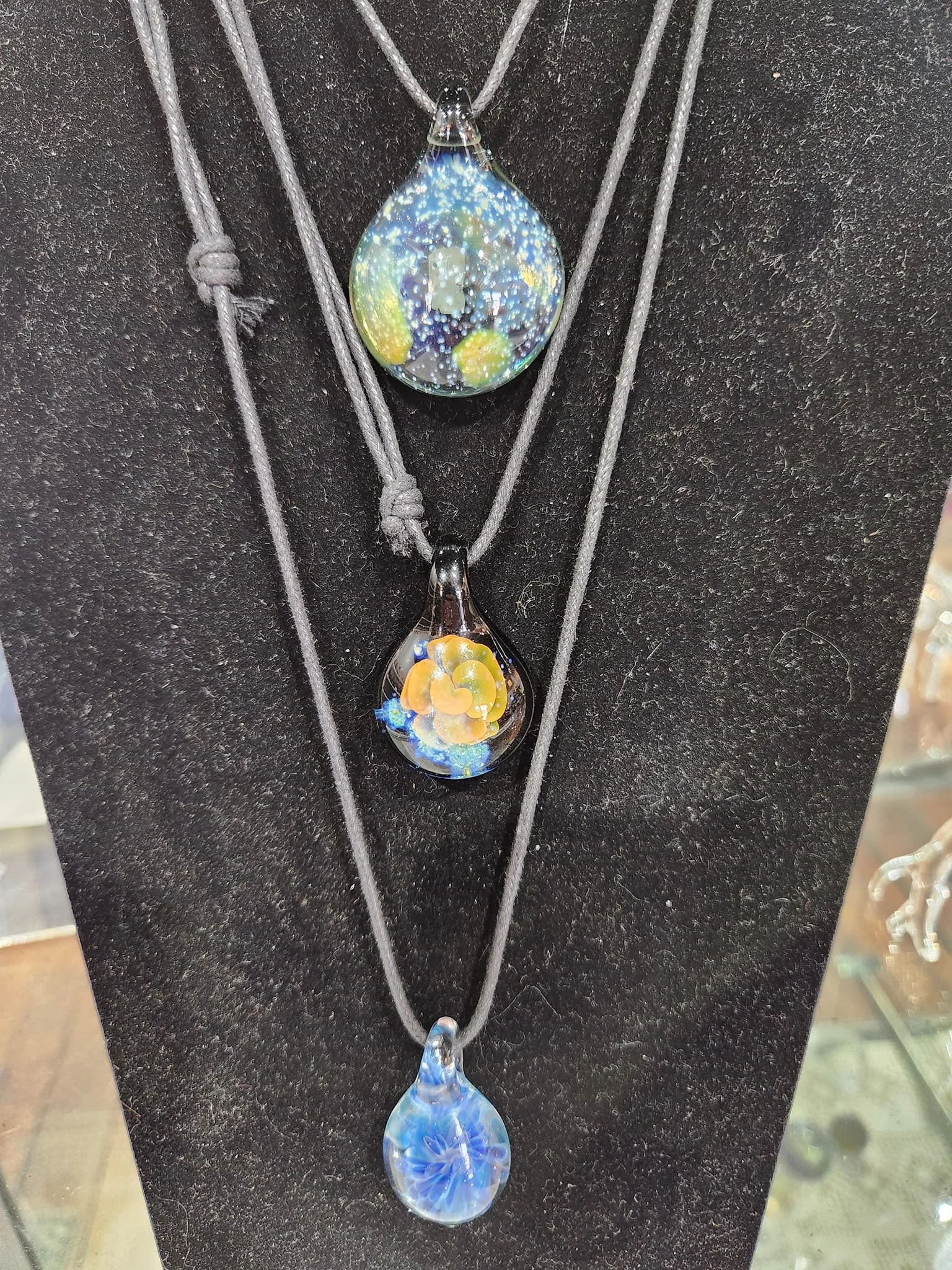Three glass pendants with colorful, abstract designs hanging on gray cord necklaces displayed against a black background.