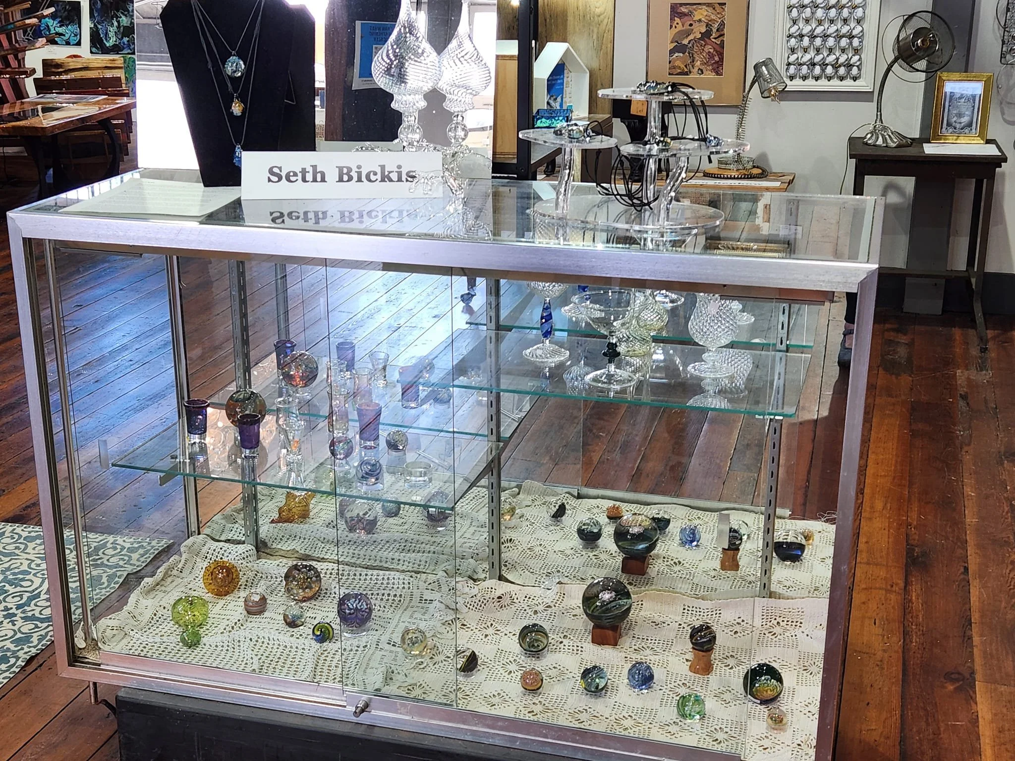 Glass display case containing multicolored marbles and glass figurines, with a sign reading "Seth Bickis." Various glassware and jewelry are on top of the case, and art and decorative items are visible in the background.