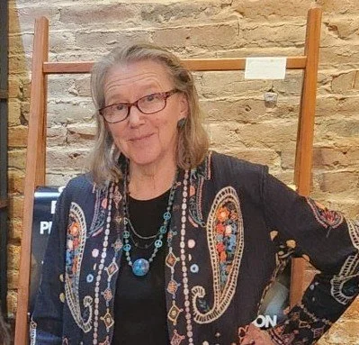 Older woman with glasses and gray hair, wearing a colorful patterned shawl and jewelry, standing in front of a brick wall and wooden ladder.