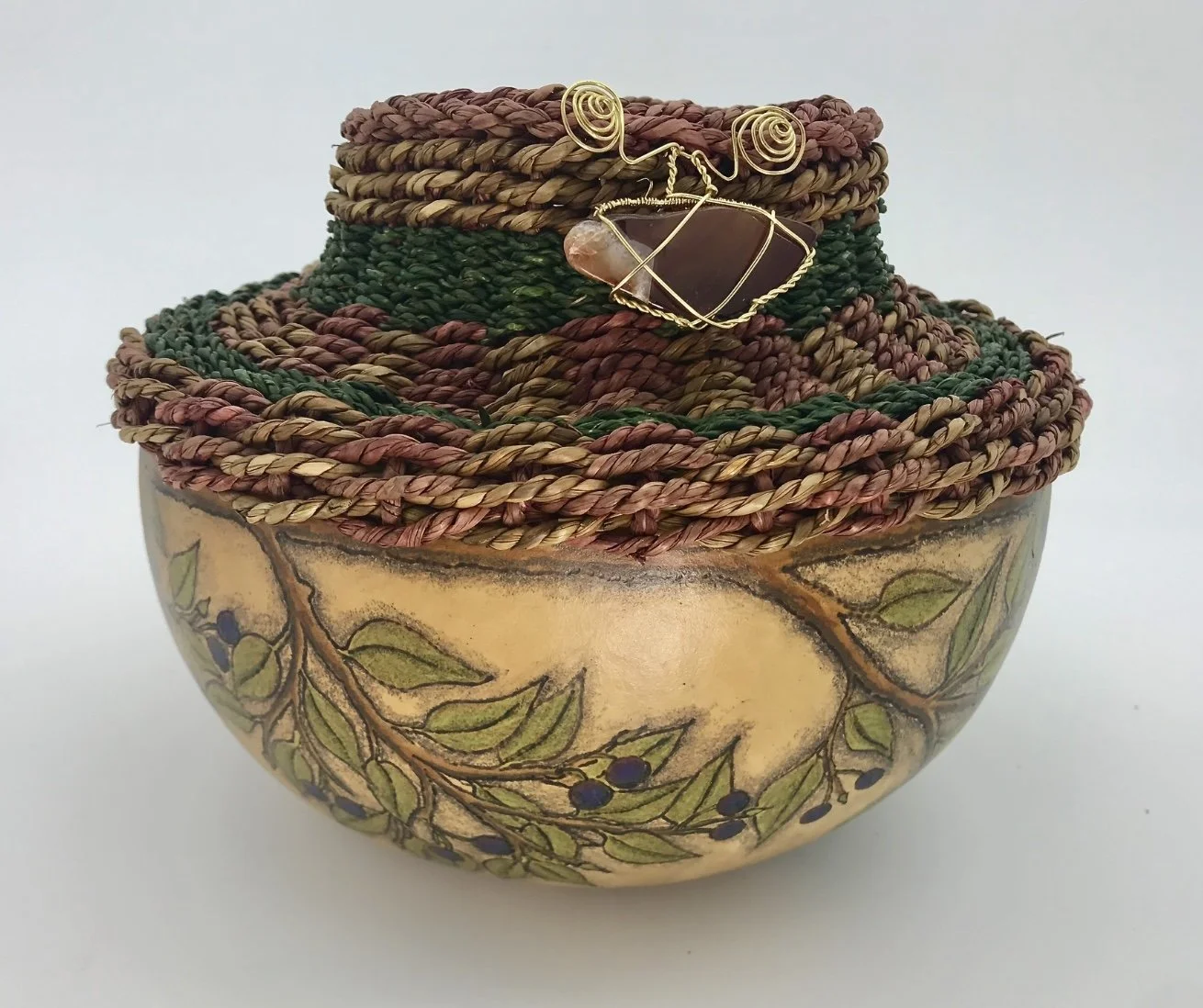 Decorative ceramic bowl with a painted branch and leaf design, filled with colorful woven fiber jewelry, including a wire-wrapped stone pendant.