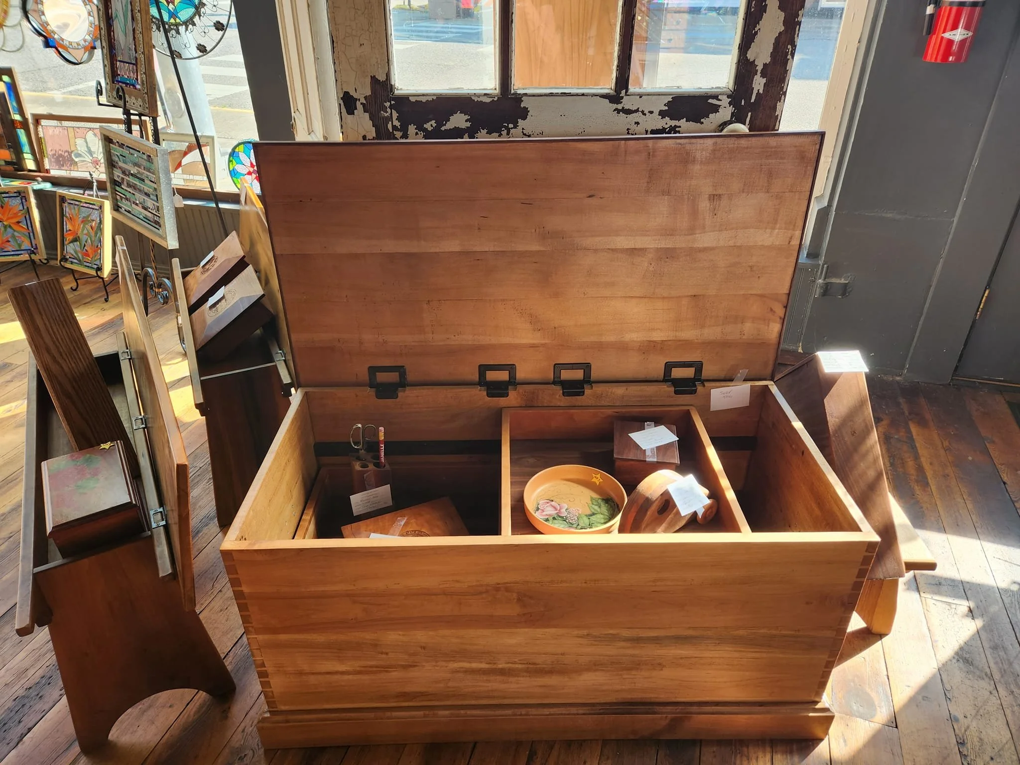 Empty wooden chest with an open lid inside an art store, with various decorative items and paintings displayed nearby, sunlight streaming in through a window.