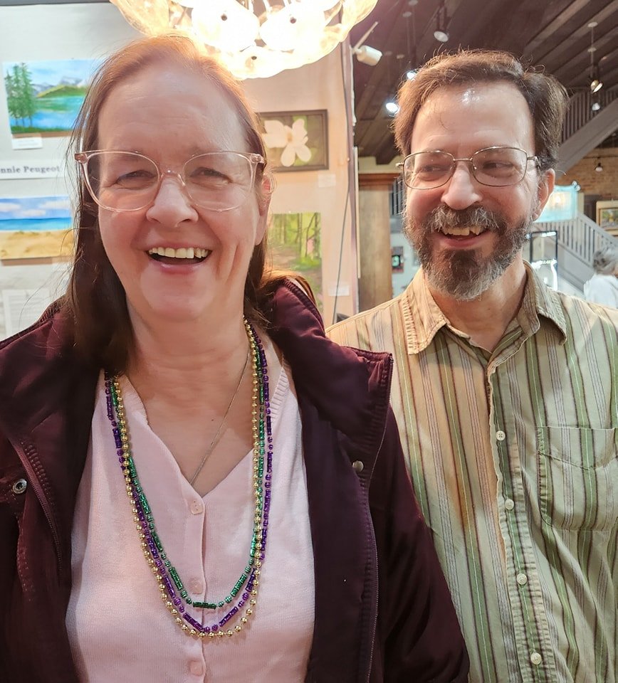 A smiling woman with glasses and layered beaded necklaces standing next to a smiling man with glasses and a striped shirt, indoors with artworks on the wall and a chandelier overhead.