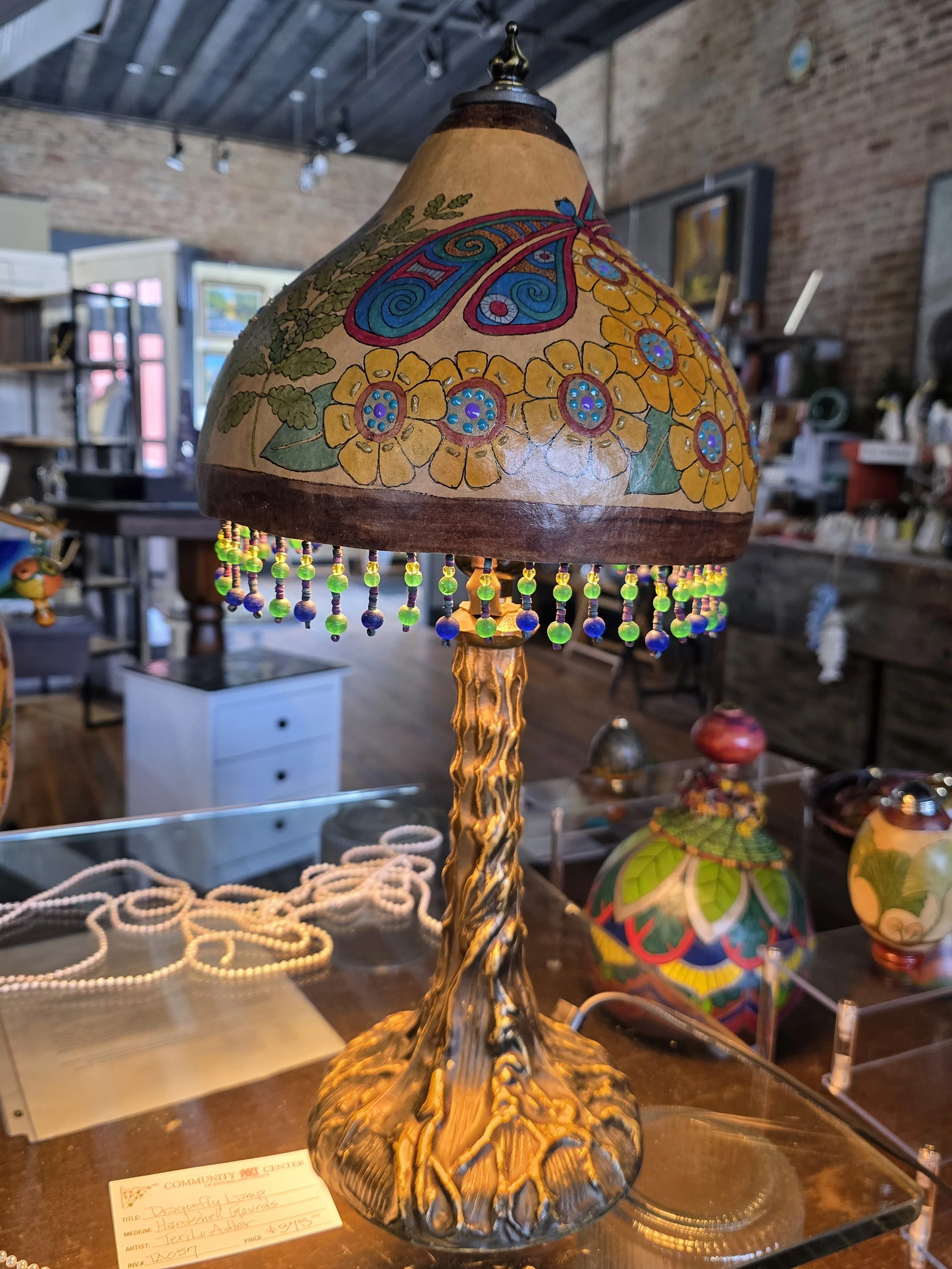 Decorative table lamp with a floral design painted on the shade, hanging beads, and a textured, twisted base. The lamp is displayed indoors in an art or craft shop.