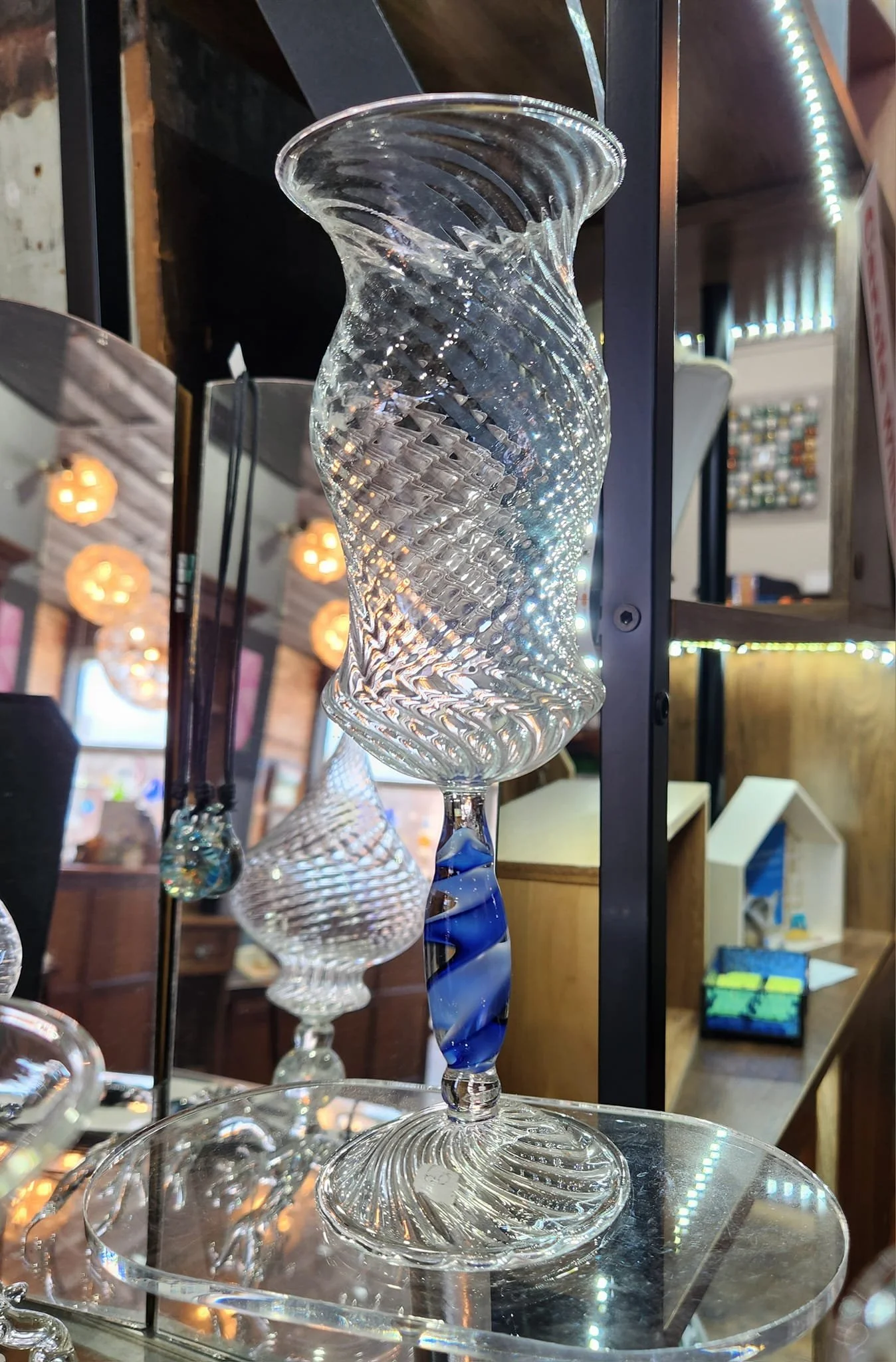 A clear, ornate glass vase with a twist pattern, displayed on a glass surface inside a shop. The vase has a wide, flared top and a twisted, colored stem.