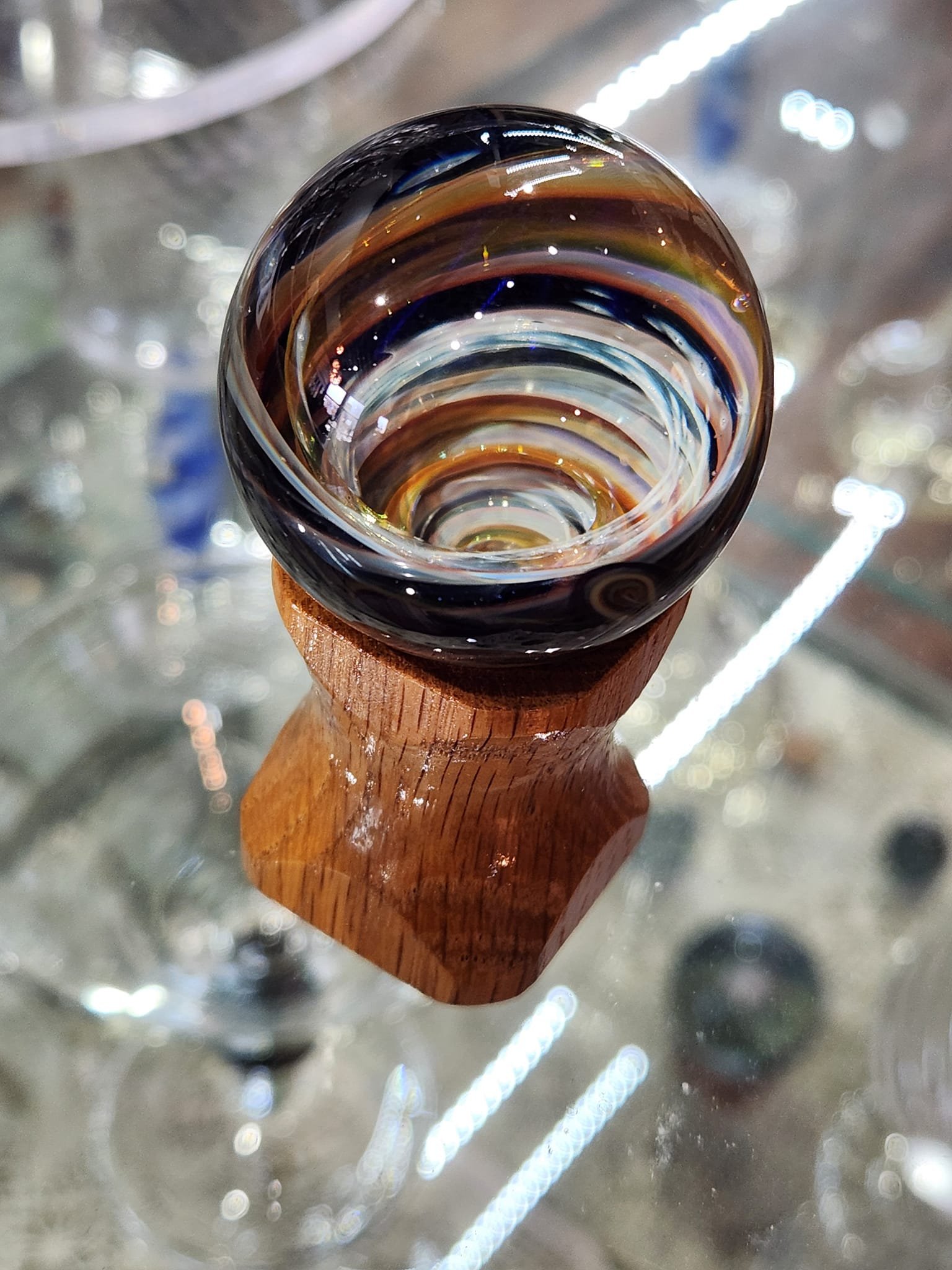Close-up of a multicolored glass swirl art piece placed on a wooden stand, viewed from above, with reflections and lights on a glass surface.