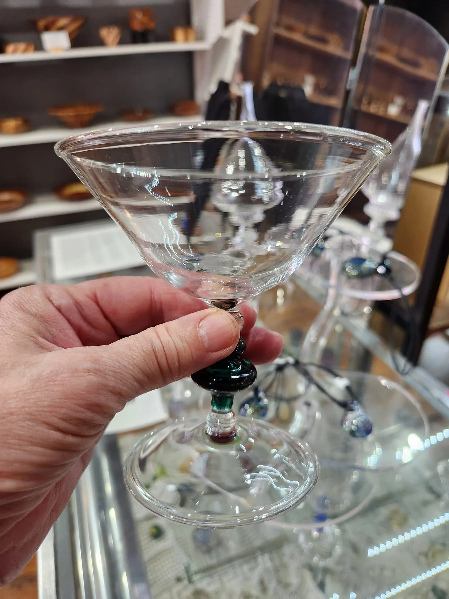 A clear glass martini glass with a decorative dark green and black accent on the stem, held by a person's hand. In the background, there are shelves with other similar glasses and various items.