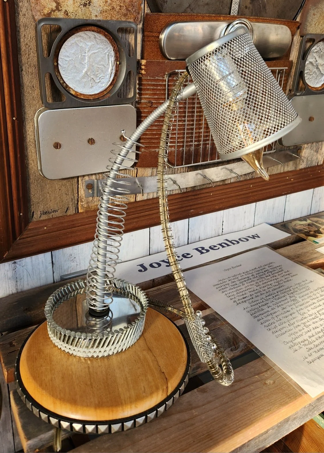 Metal lamp made from what appears to be various recycled metal parts, placed on a wooden drum and positioned in front of a wooden wall display with a written biography of Joye Benbow.